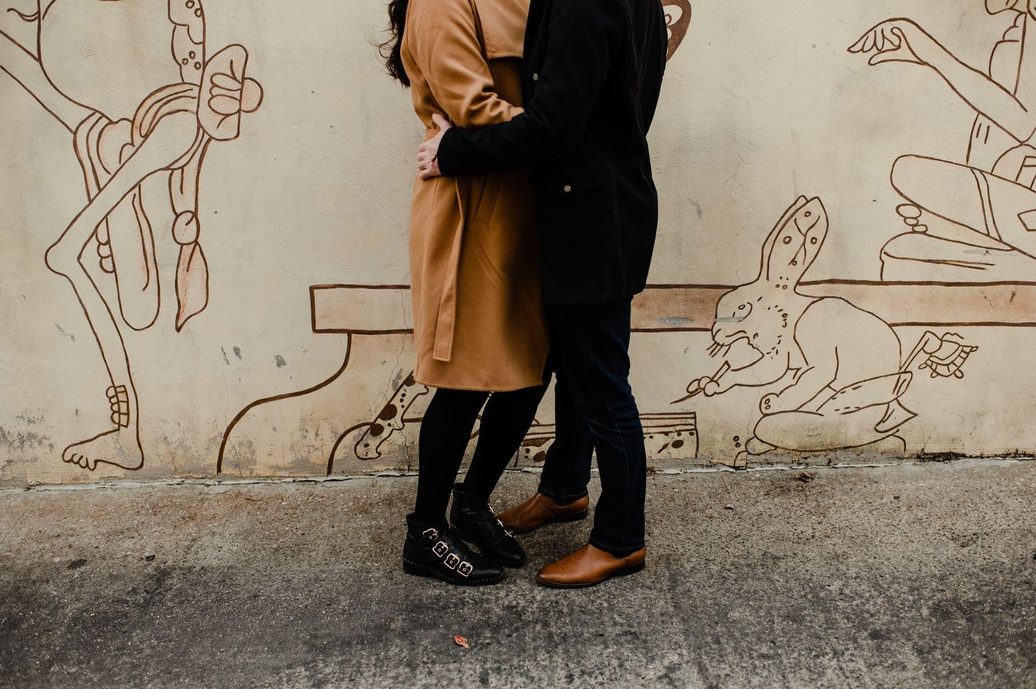 Couple embracing in front of a wall mural with line art drawings, wearing black and tan outfits.