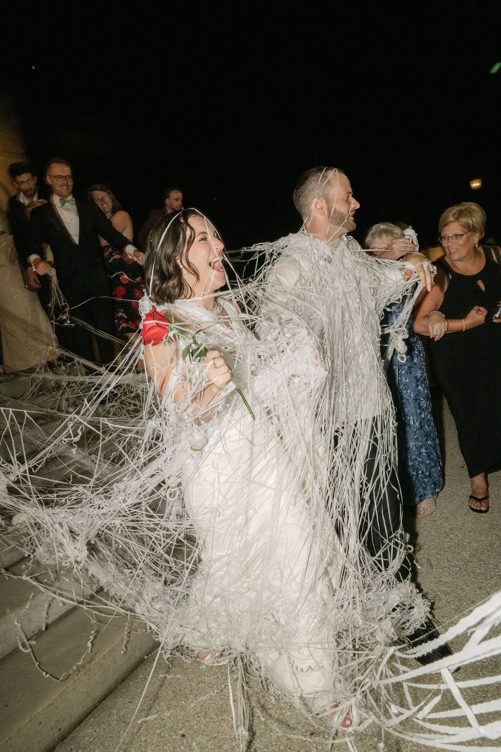 Wedding couple gets wrapped in toilet paper during fun reception game at nighttime celebration.