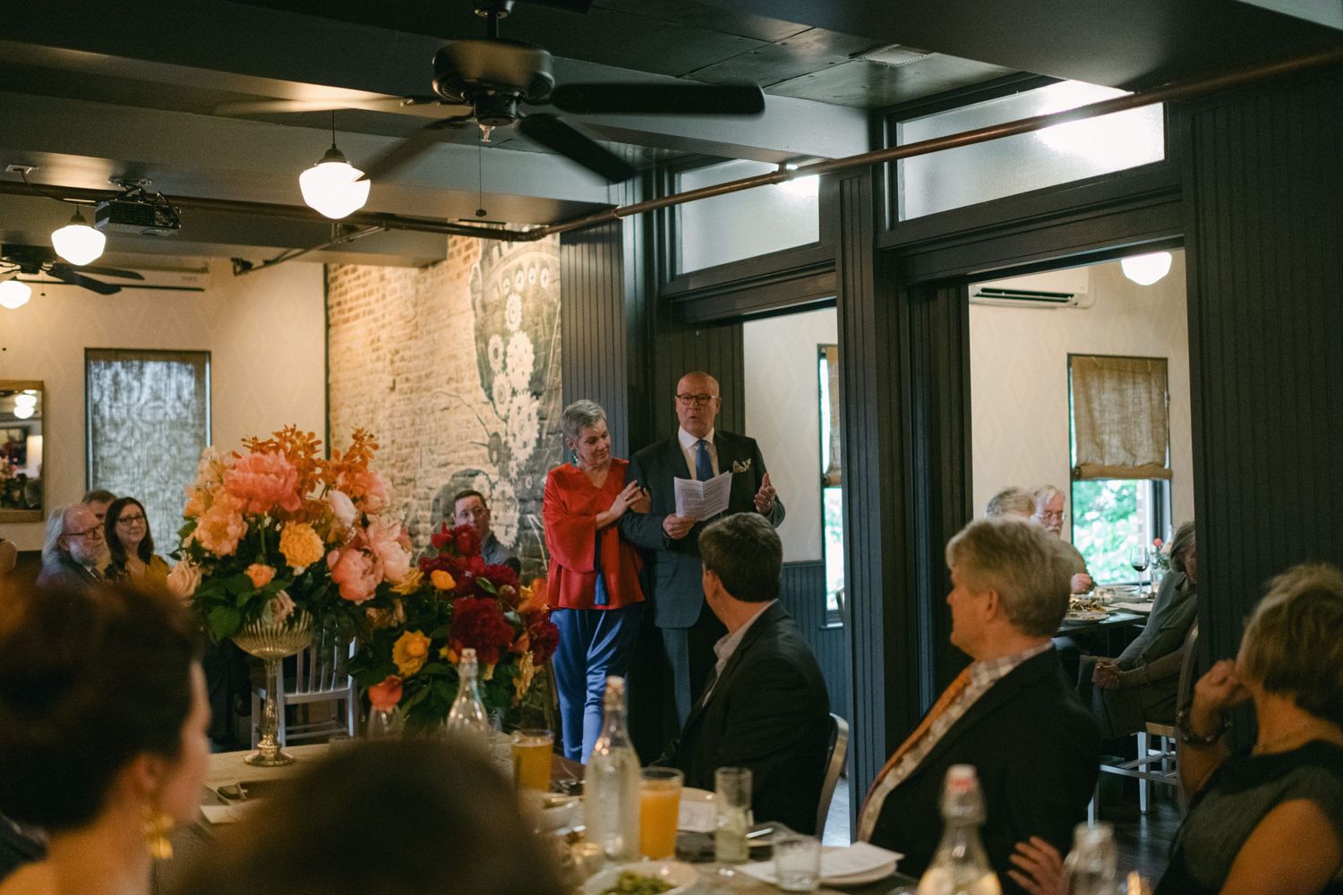 Wedding reception guests gather in a cozy restaurant space with exposed brick walls and ceiling fans.