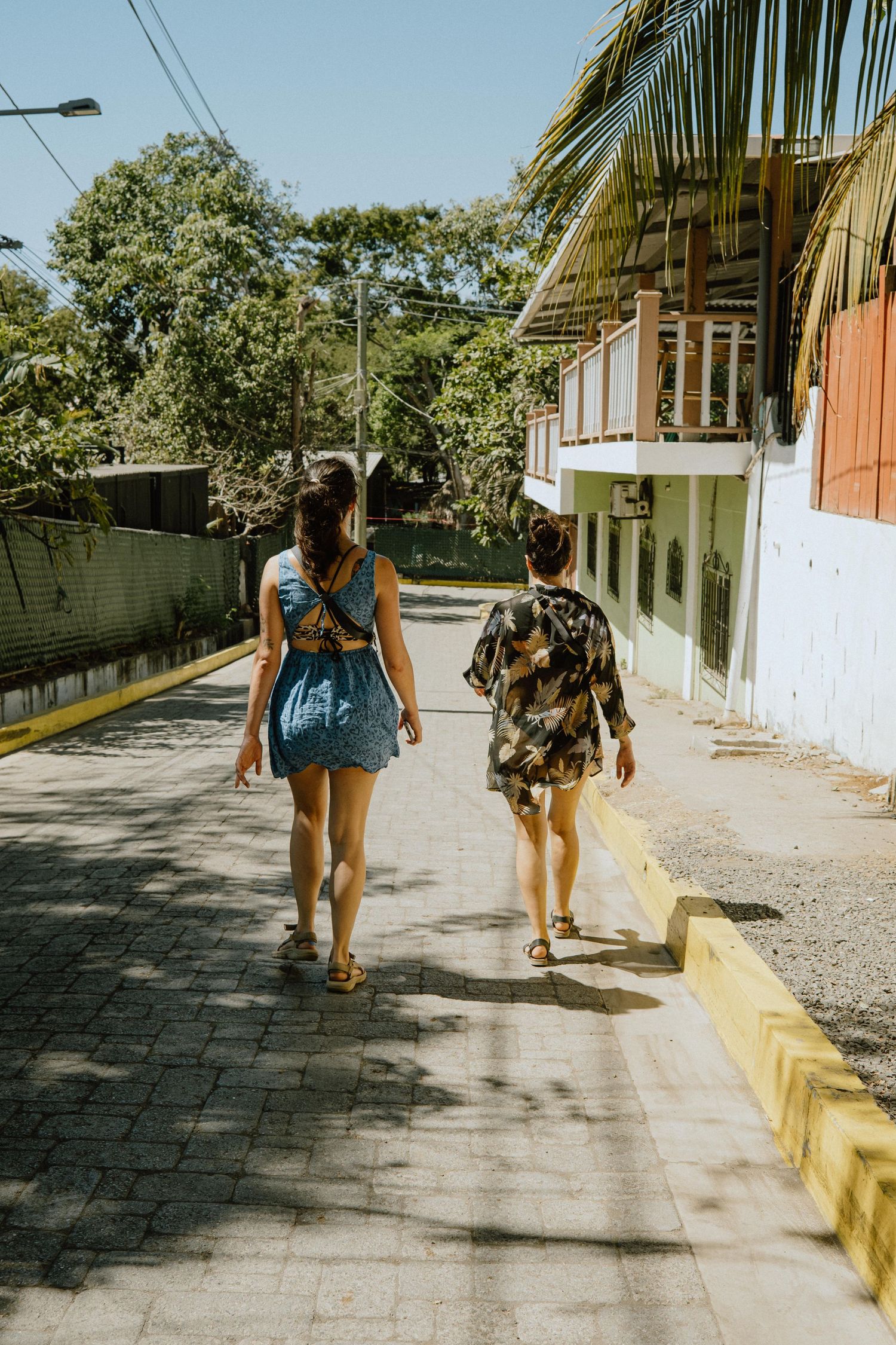 Two people walking down a sunny tropical street lined with palm trees and buildings.
