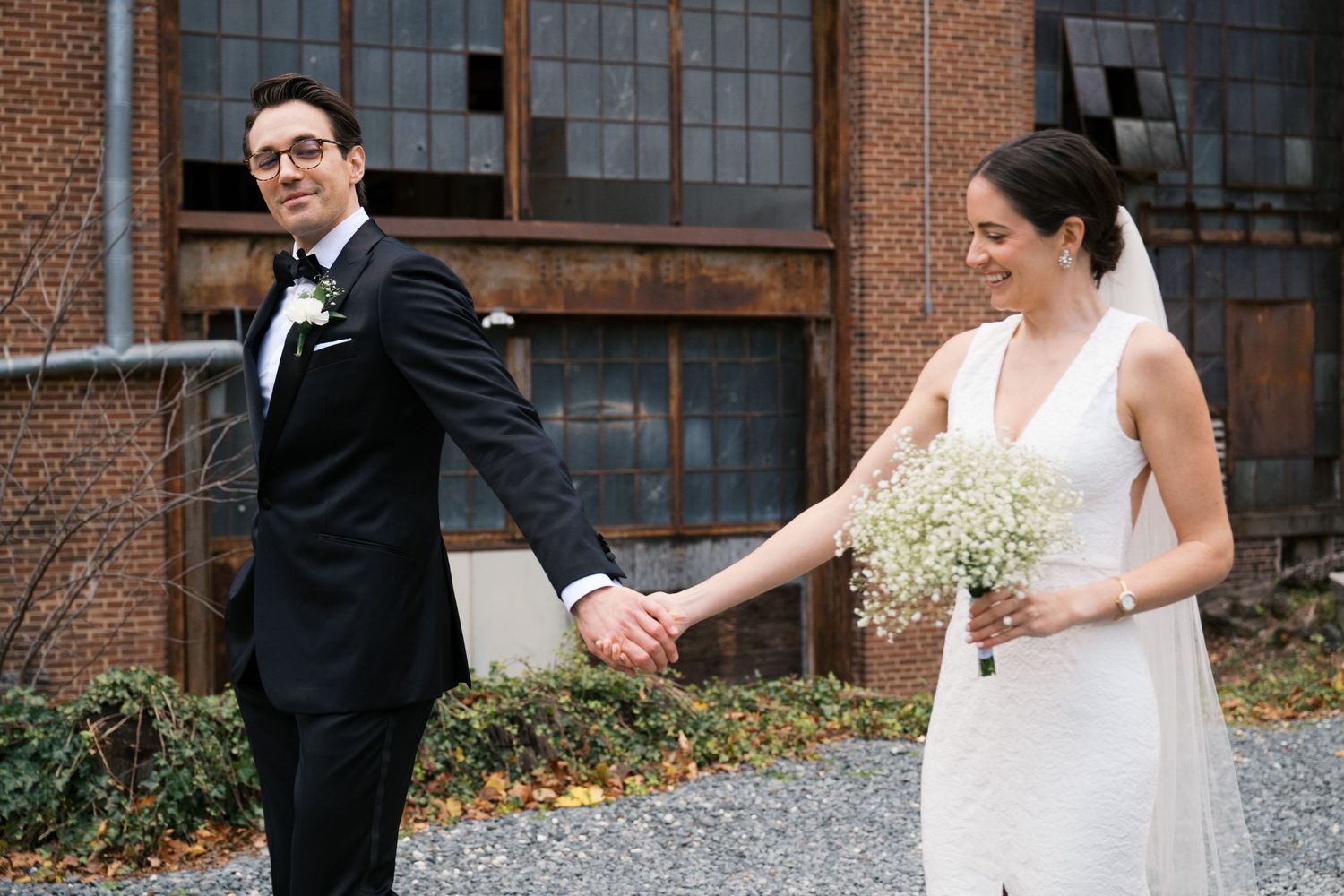 A bride in white dress holds baby's breath bouquet walking with groom by brick building.