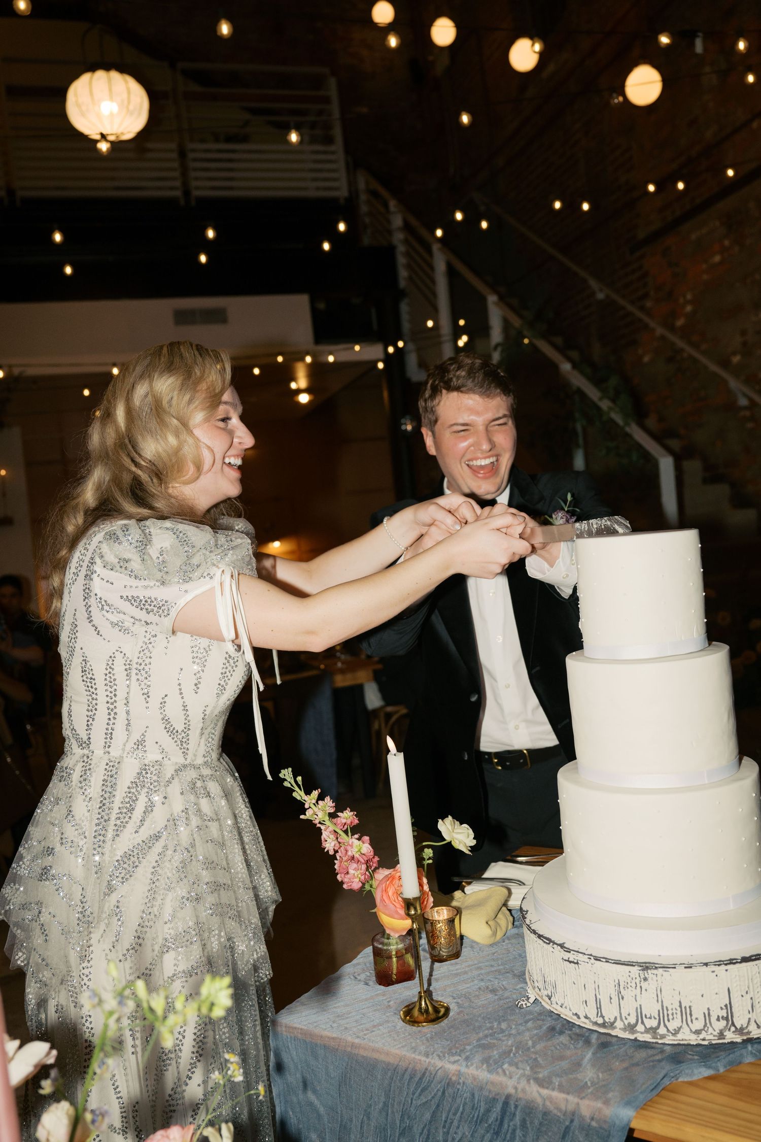 A couple cuts their elegant white wedding cake together under hanging lights.