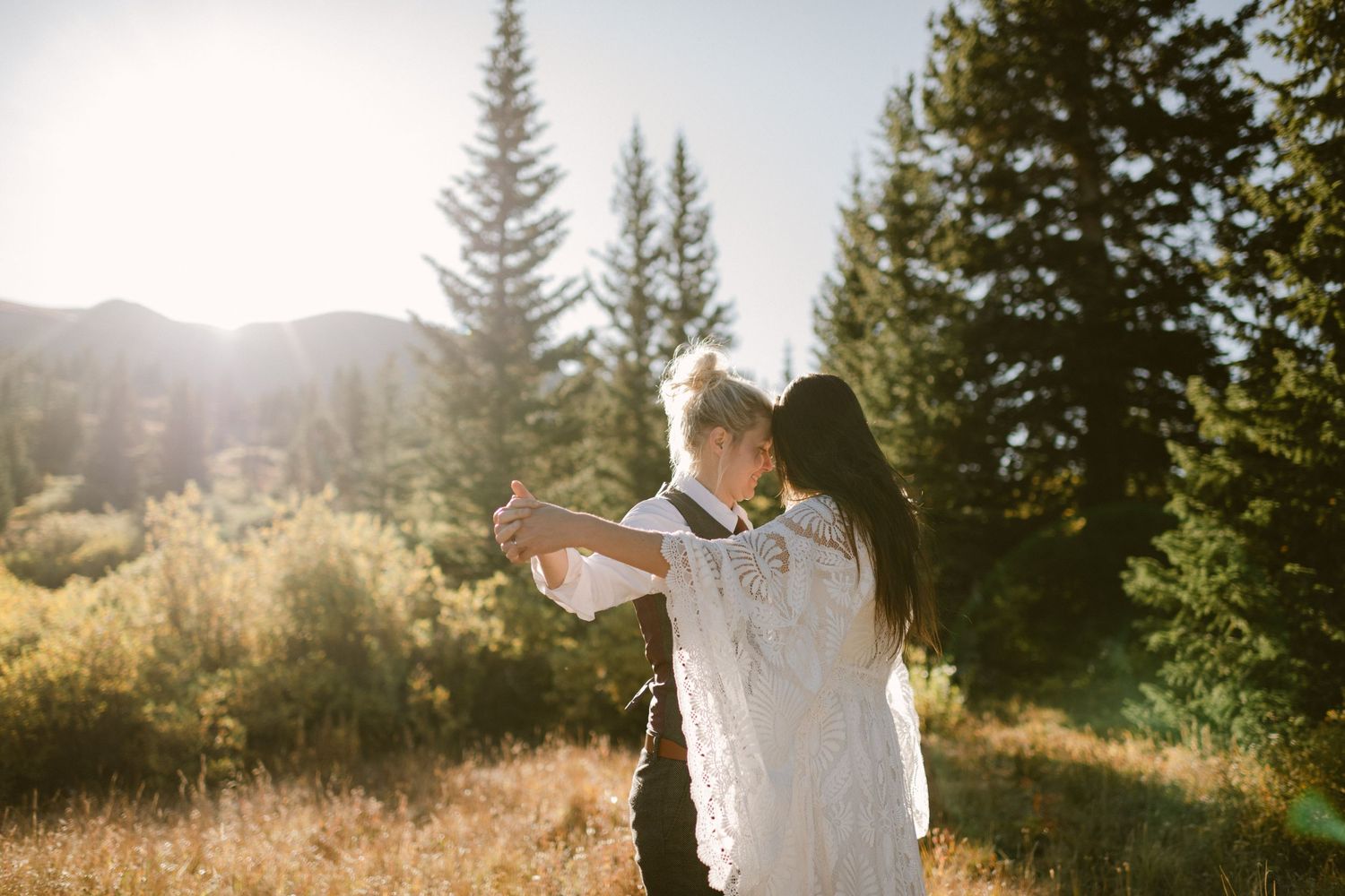 A couple embracing in front of sunlit mountains and pine trees.