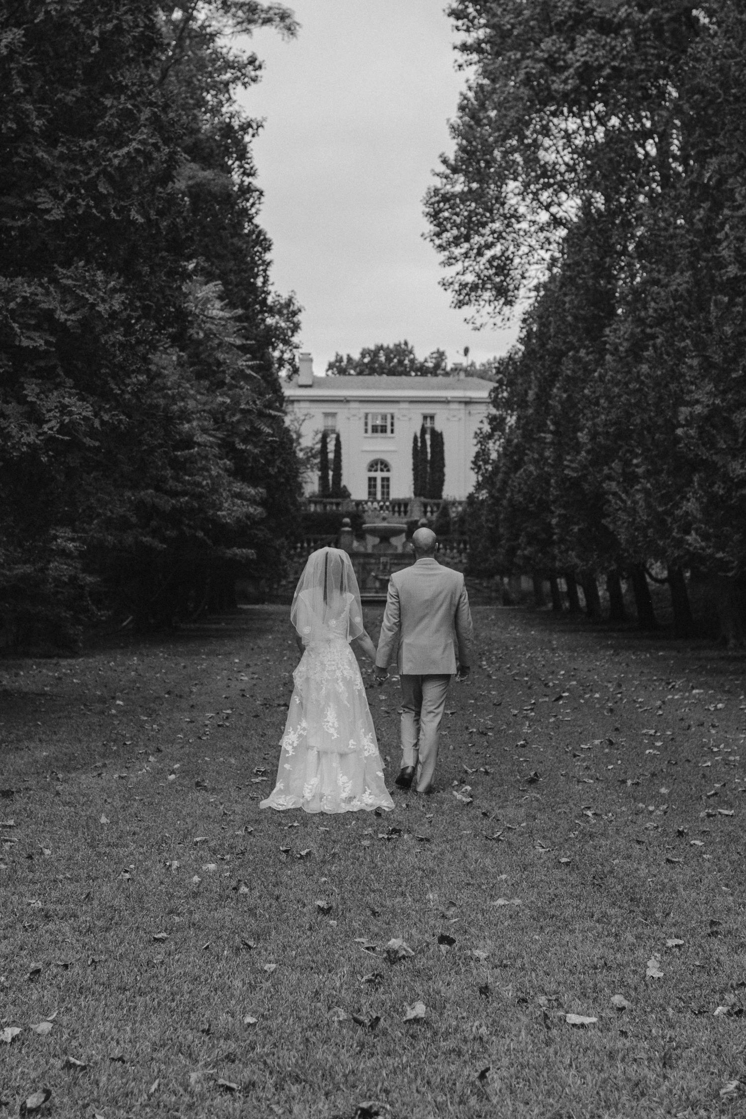 Black and white photo of couple walking toward a grand estate through a tree-lined path.