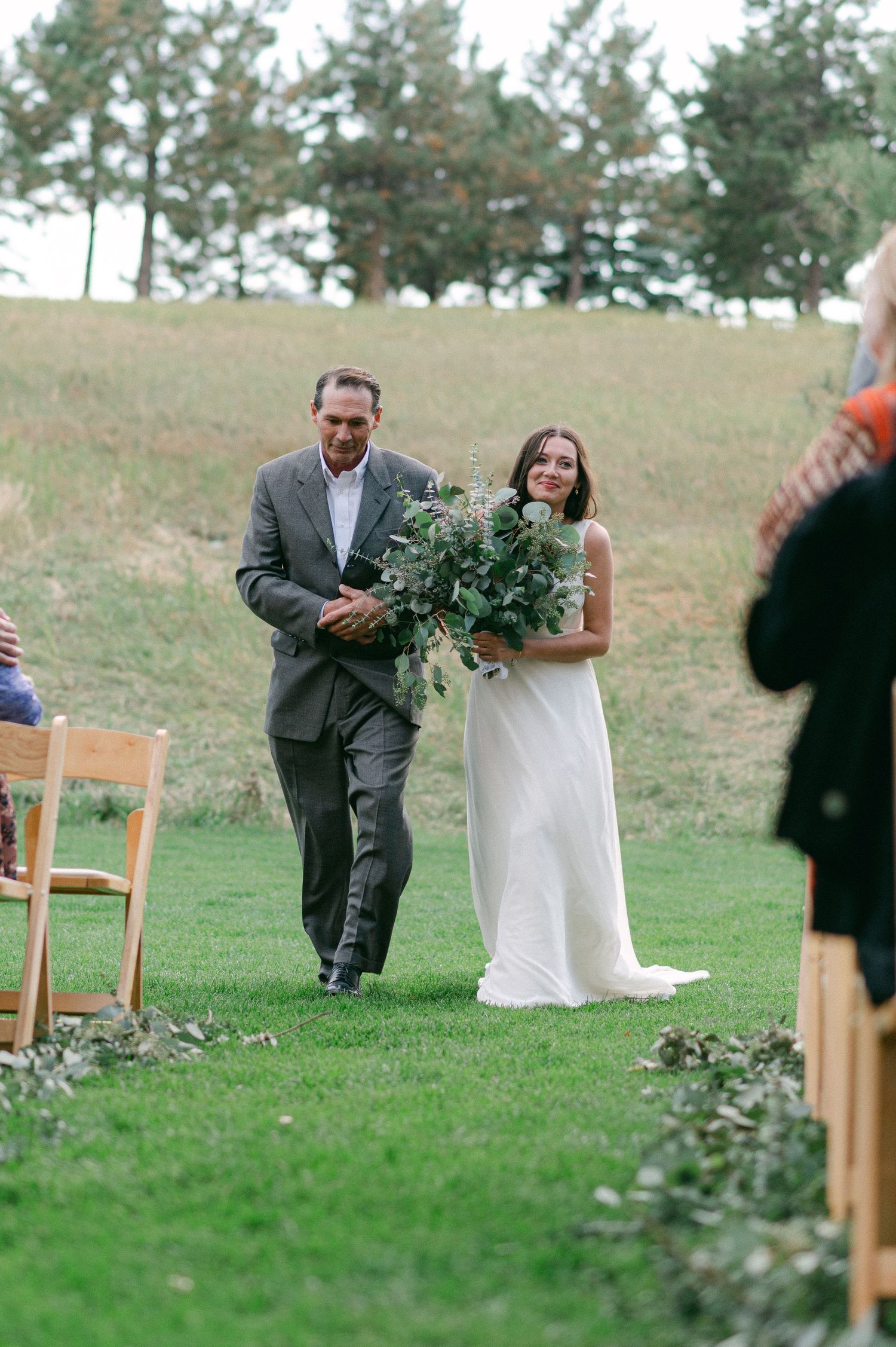 Wedding processional on a grassy hillside with pine trees in the background.