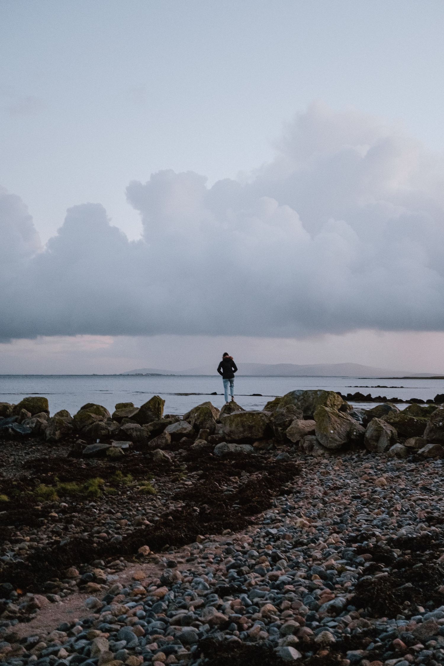 A solitary figure stands on rocky shoreline overlooking the ocean at dusk.