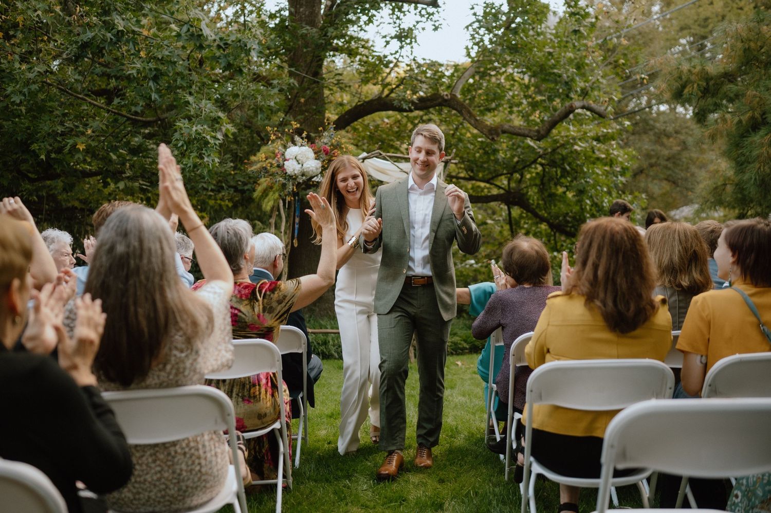 Wedding guests cheer as newlyweds walk down outdoor garden aisle surrounded by white chairs.
