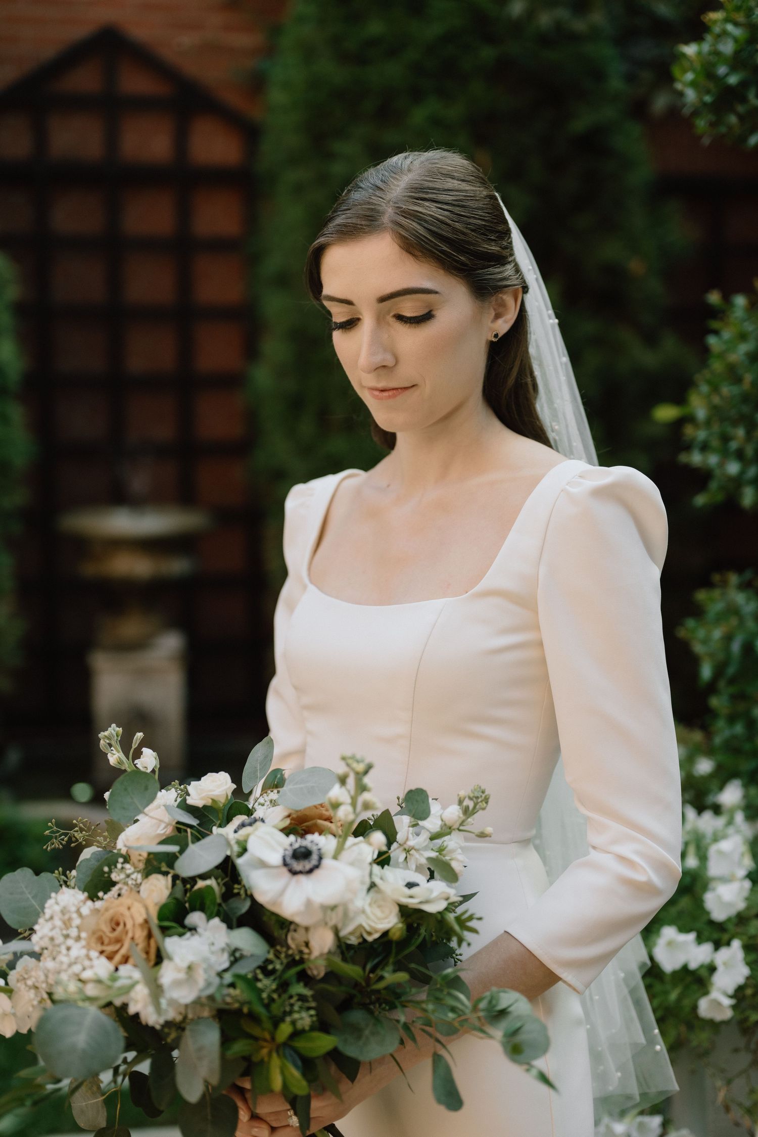 A bride in an elegant long-sleeved white dress holds a lush bouquet of flowers with eucalyptus against a garden backdrop.