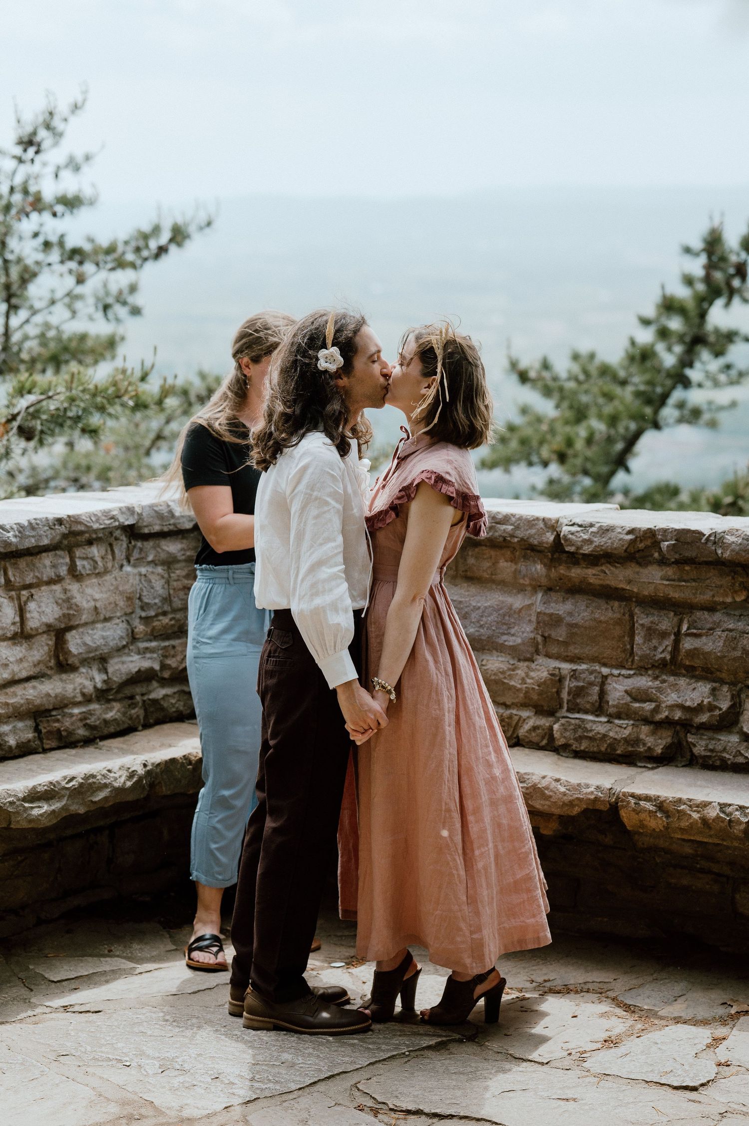 Three people embracing at a scenic mountain overlook with stone walls.