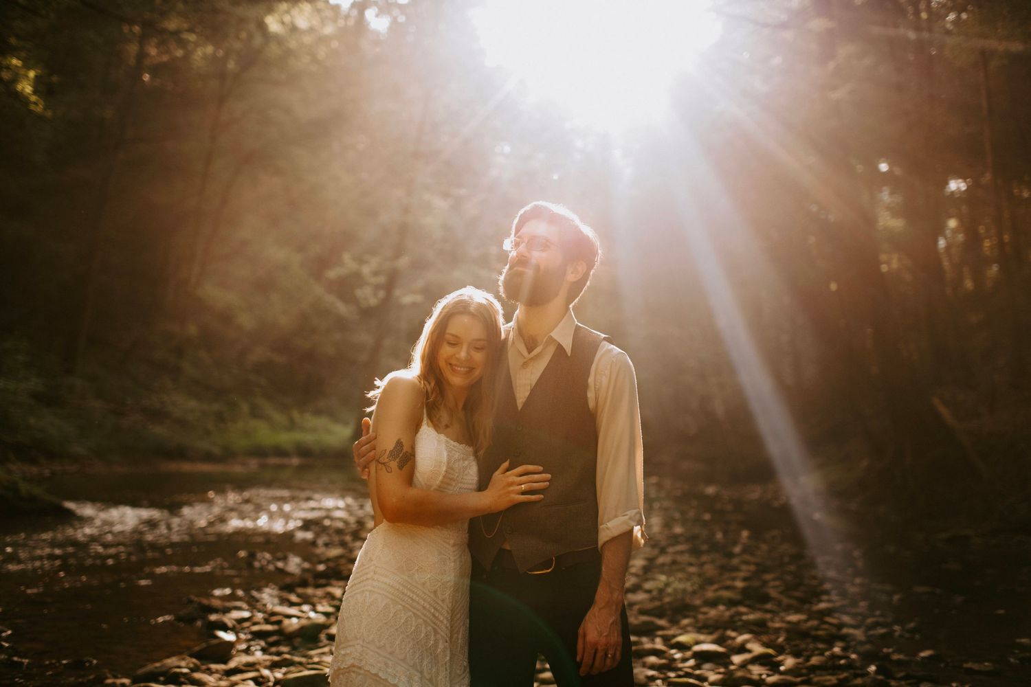 Romantic couple embraces in sunlit forest setting with golden rays streaming through trees.