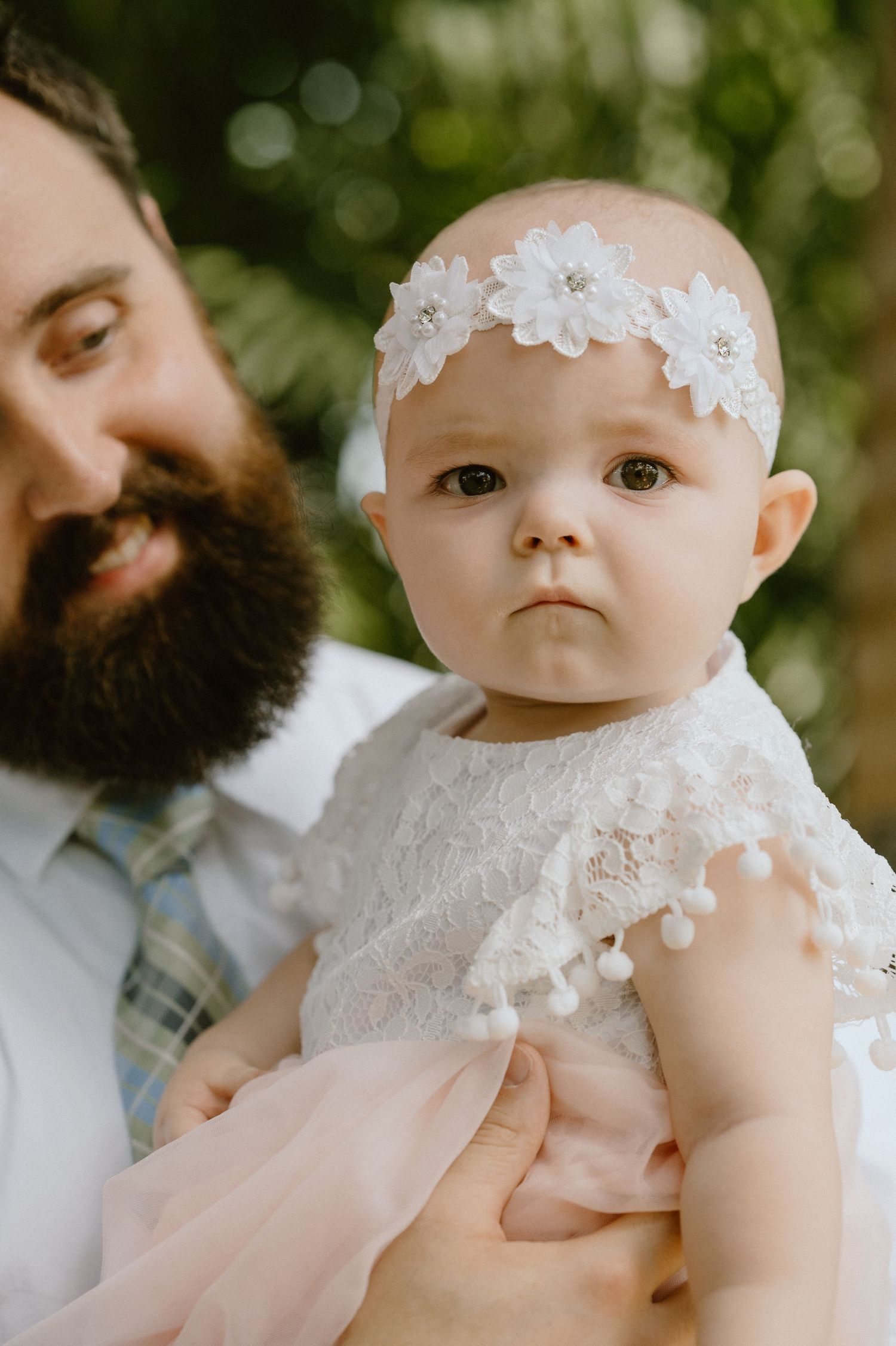 A baby wearing a delicate white dress and floral headband is held against a natural outdoor background.