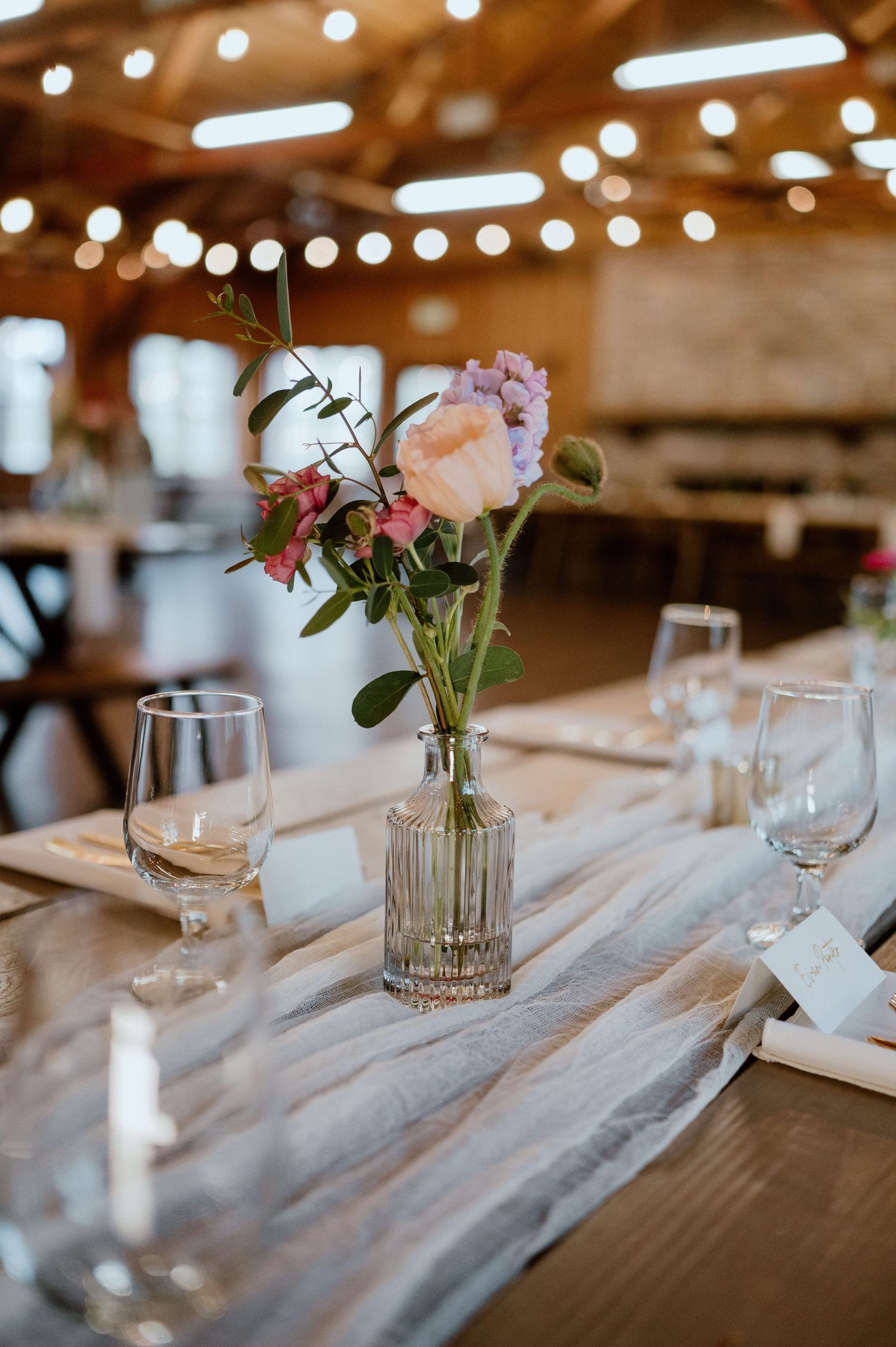 An elegant table setting with pink roses in a glass vase, wine glasses, and string lights above.