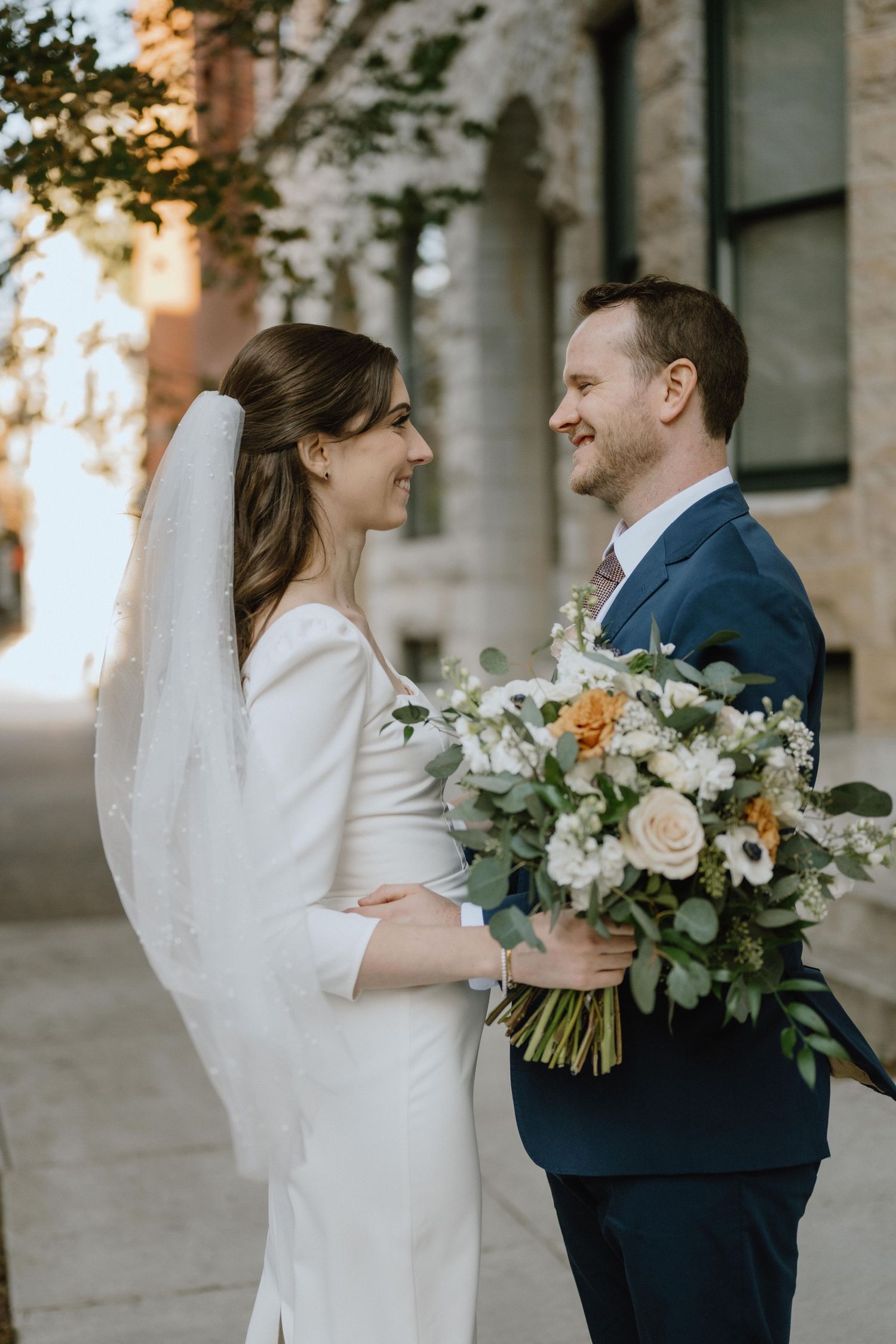 Golden hour wedding portrait with elegant bouquet against stone building backdrop.