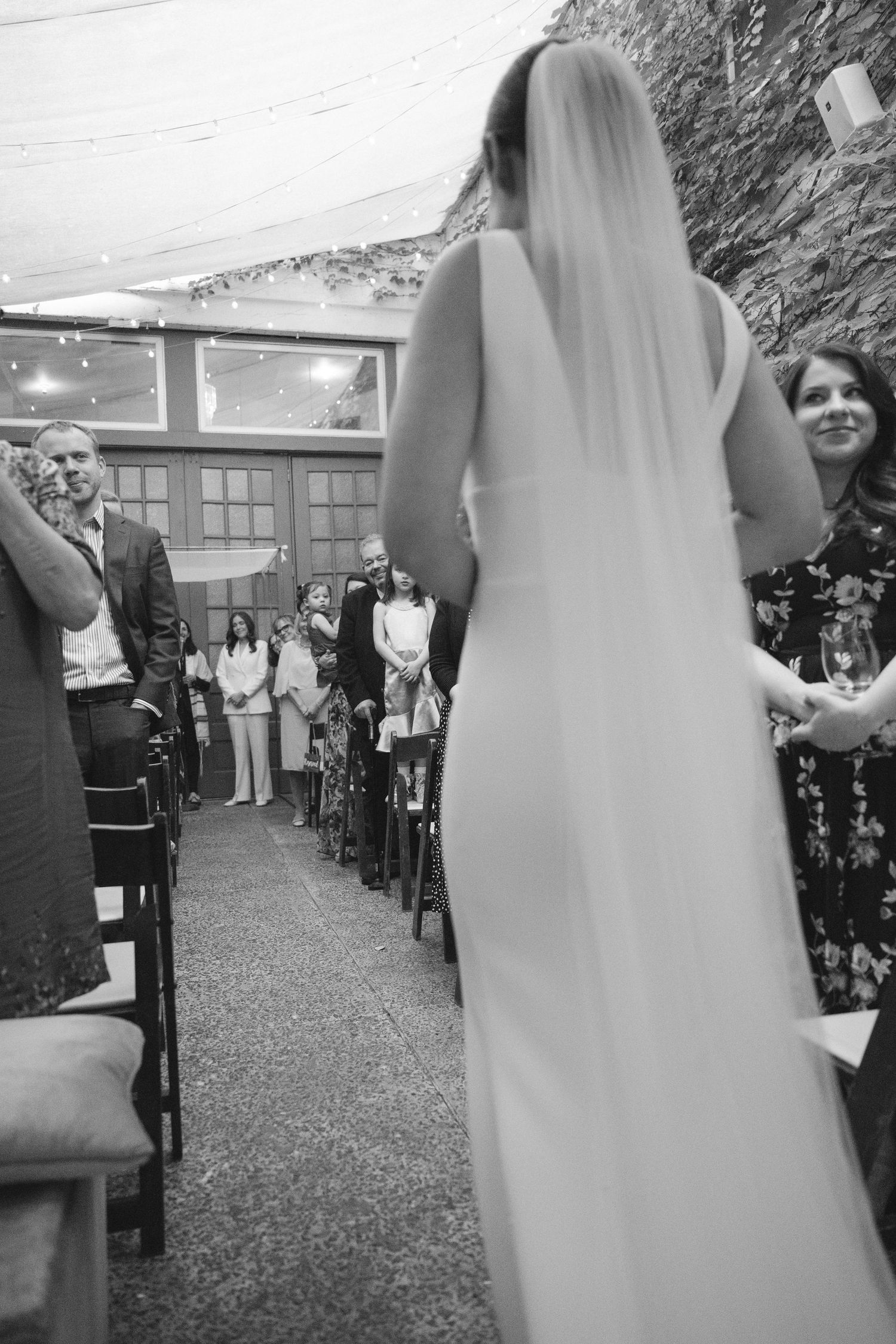 A bride in a white wedding dress stands at the front of a ceremony space while guests look on from their seats.