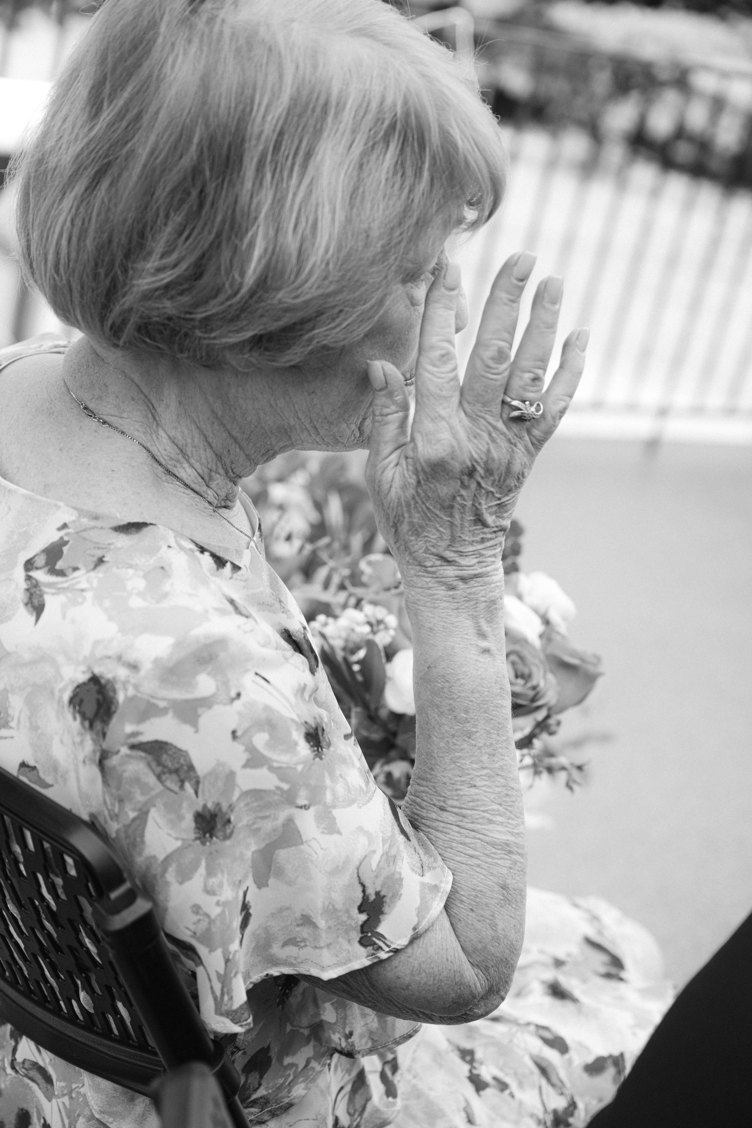 An elderly woman in a floral dress covers her mouth with her hand while wearing rings.