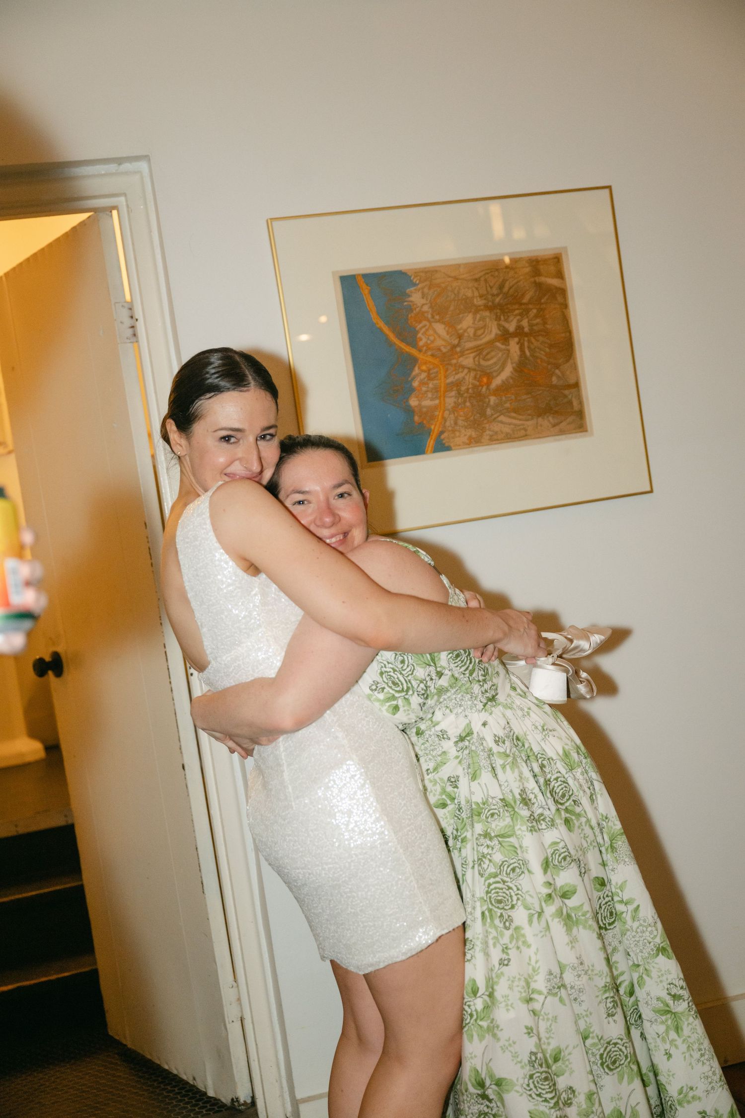 Two people embrace in a candid moment, one wearing a white lace dress and one in a floral patterned dress.