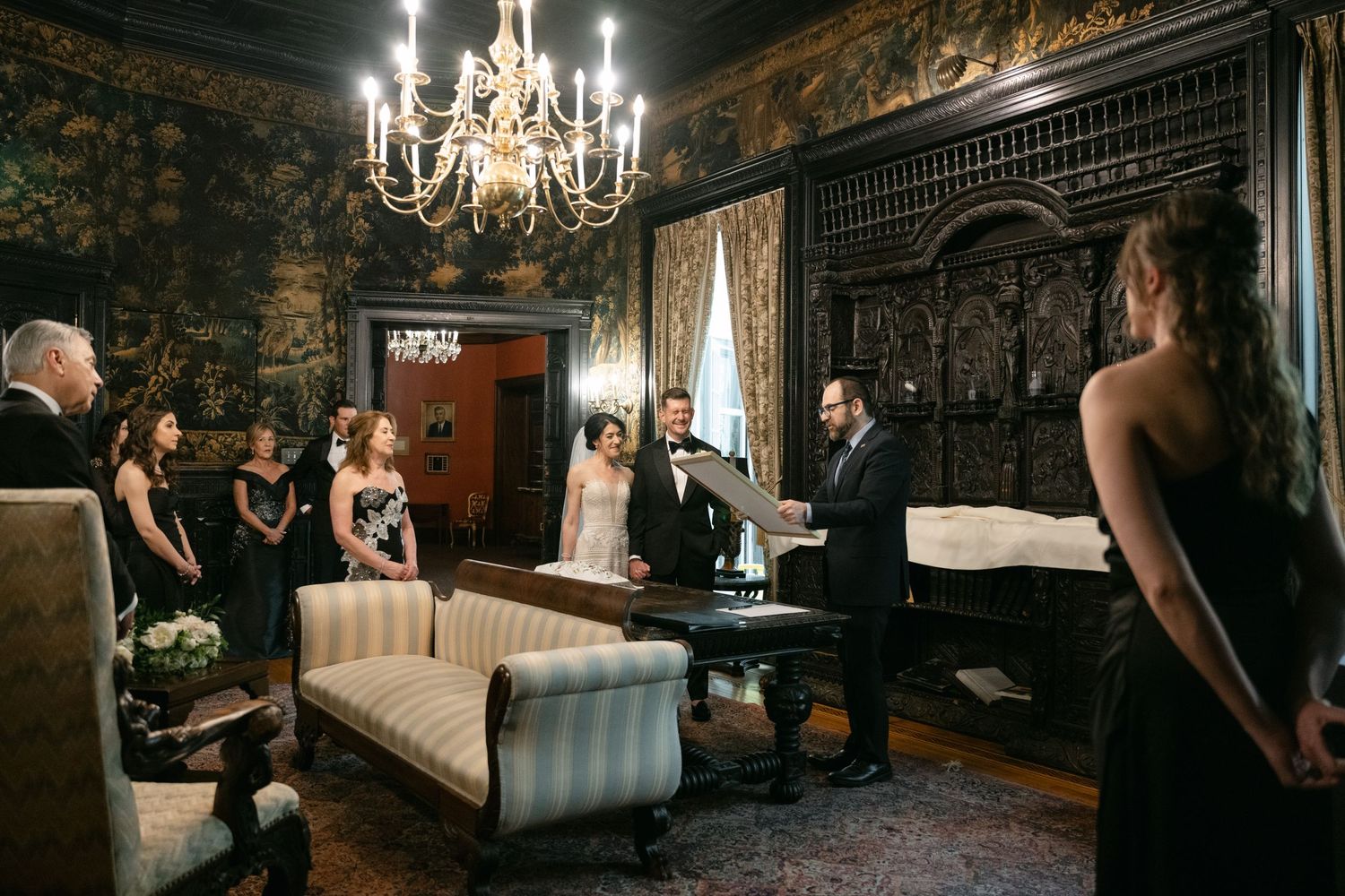 An intimate wedding ceremony takes place inside an elegant vintage room with dark walls and a crystal chandelier.