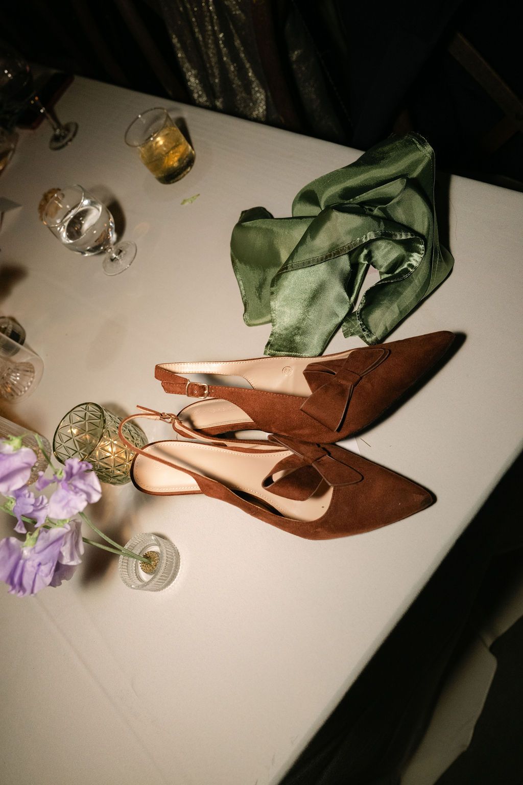 Brown dress shoes and green fabric on a white tablecloth with wine glasses and purple flowers.