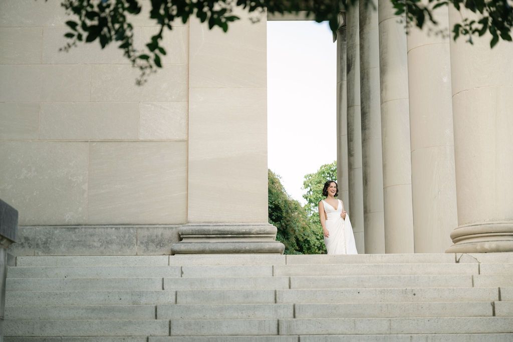 A figure in a white dress stands elegantly between classic stone columns on grand marble steps.