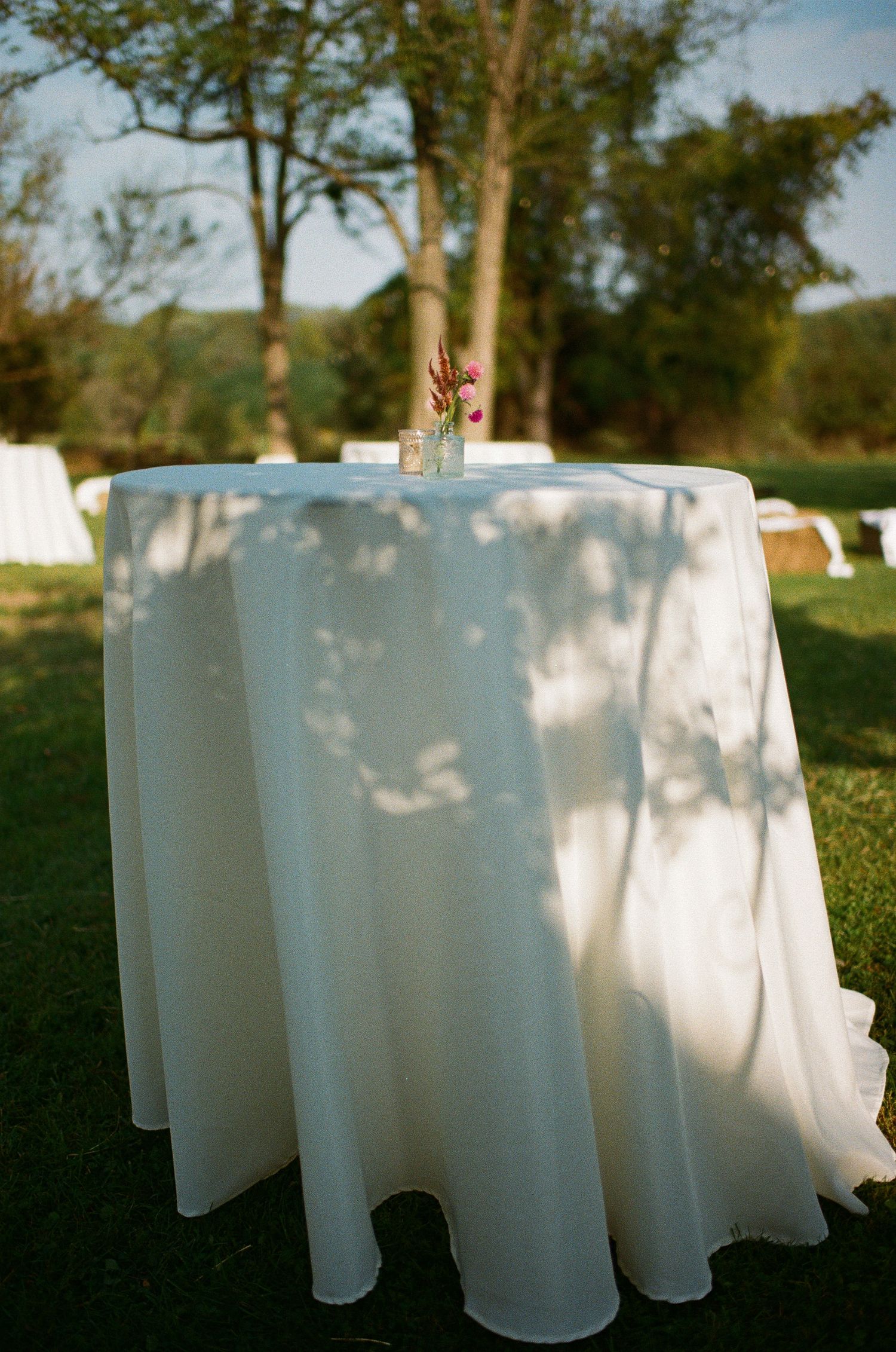 White tablecloths draped over round tables in an outdoor garden setting with dappled sunlight.