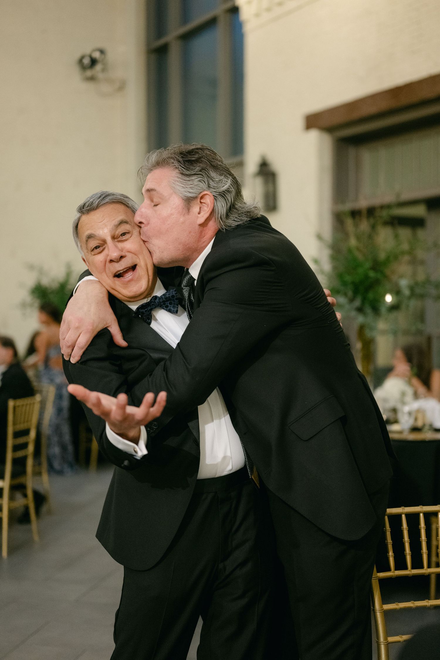 Two men in tuxedos share a playful moment at a formal reception.