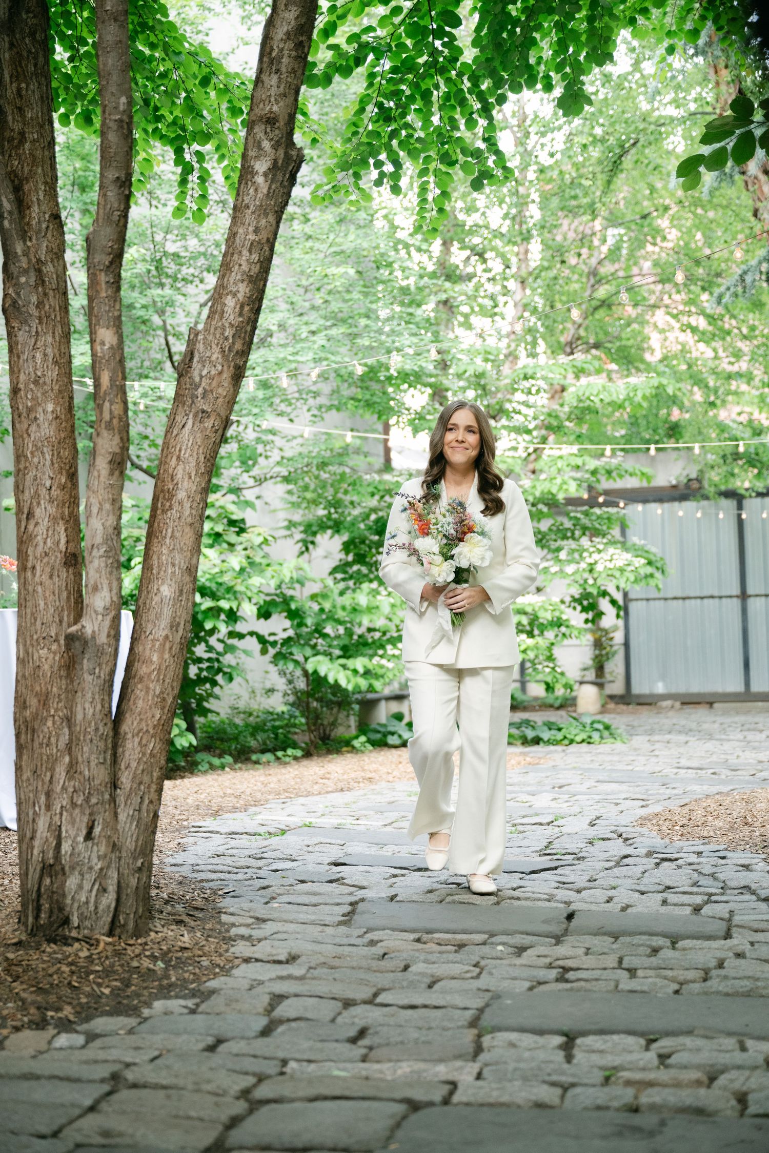 Someone in a white pantsuit walks along a cobblestone path holding a bouquet in a lush garden setting.