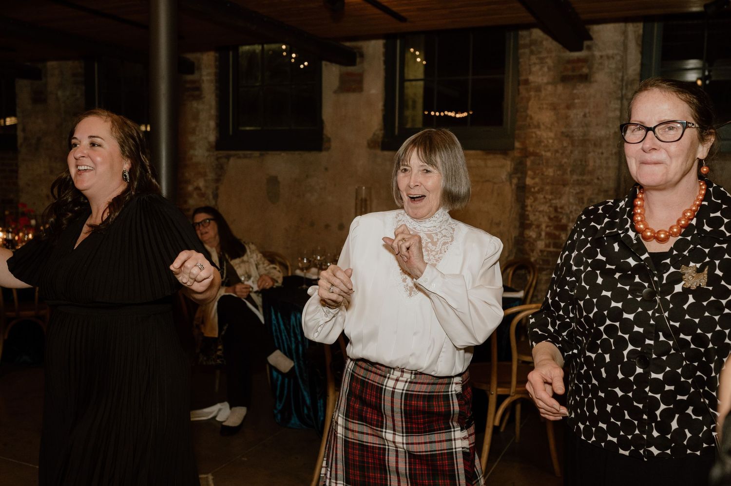 Three people dancing enthusiastically at an indoor celebration with exposed brick walls.