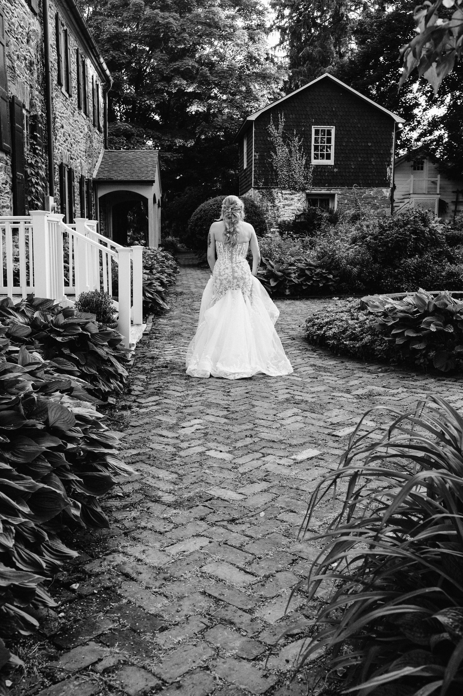 A black and white photo of a bride walking on a cobblestone path between lush garden foliage.