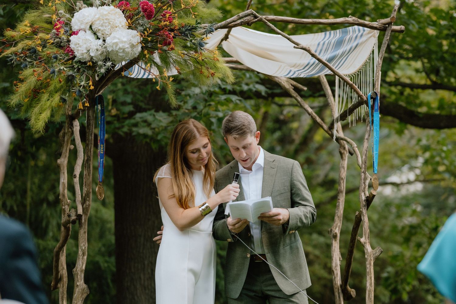 Couple exchanging vows under a rustic wooden arch decorated with white flowers.