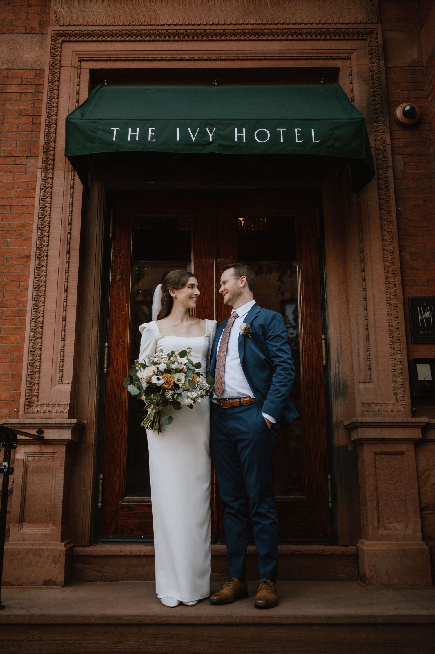 Elegant couple stands under The Ivy Hotel entrance sign in formal attire.