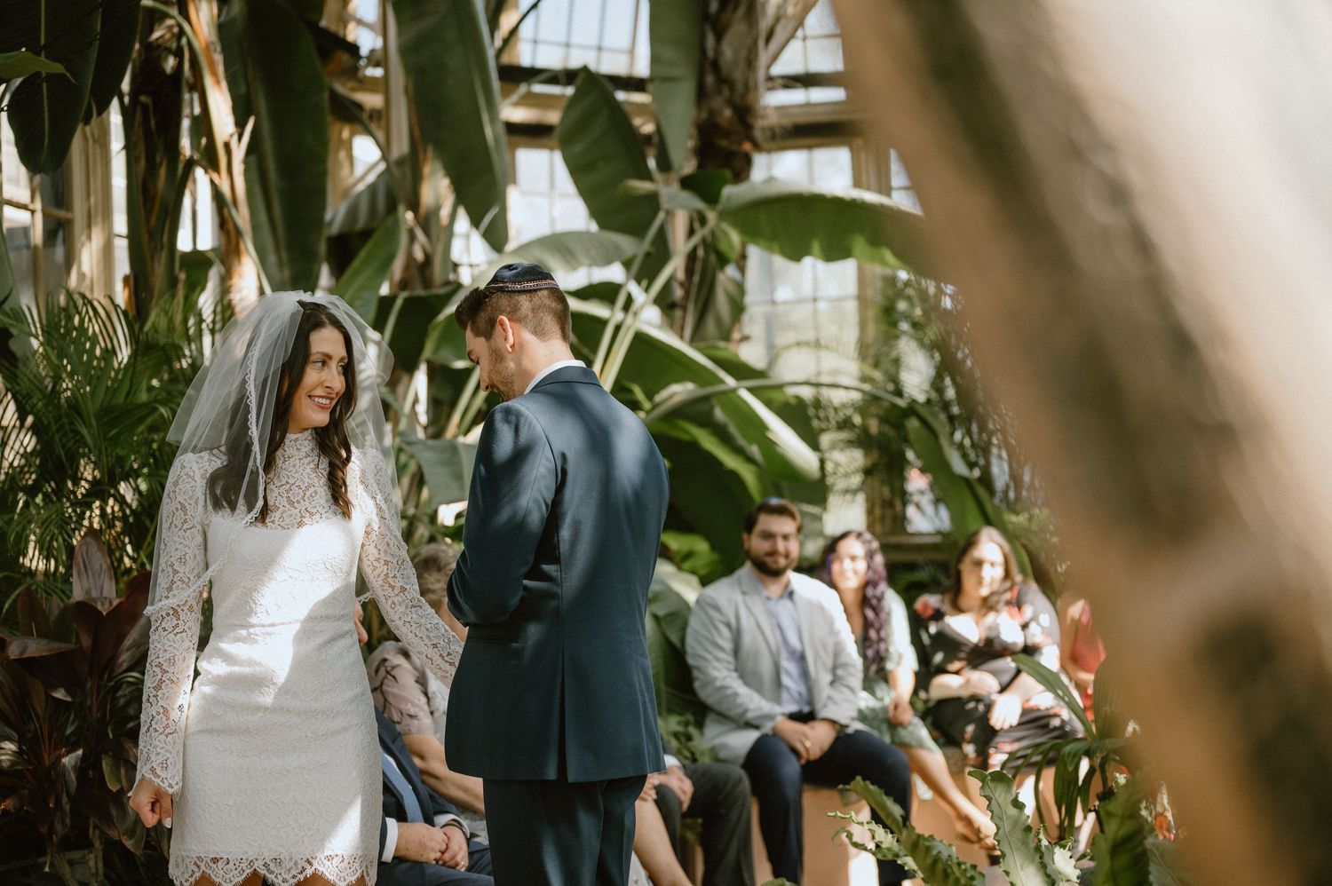 A couple shares a moment among lush greenery in a sunlit conservatory during their wedding ceremony.