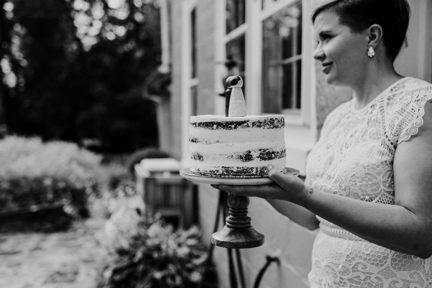 Someone in a lace dress holds a tiered white wedding cake on a decorative stand outdoors.