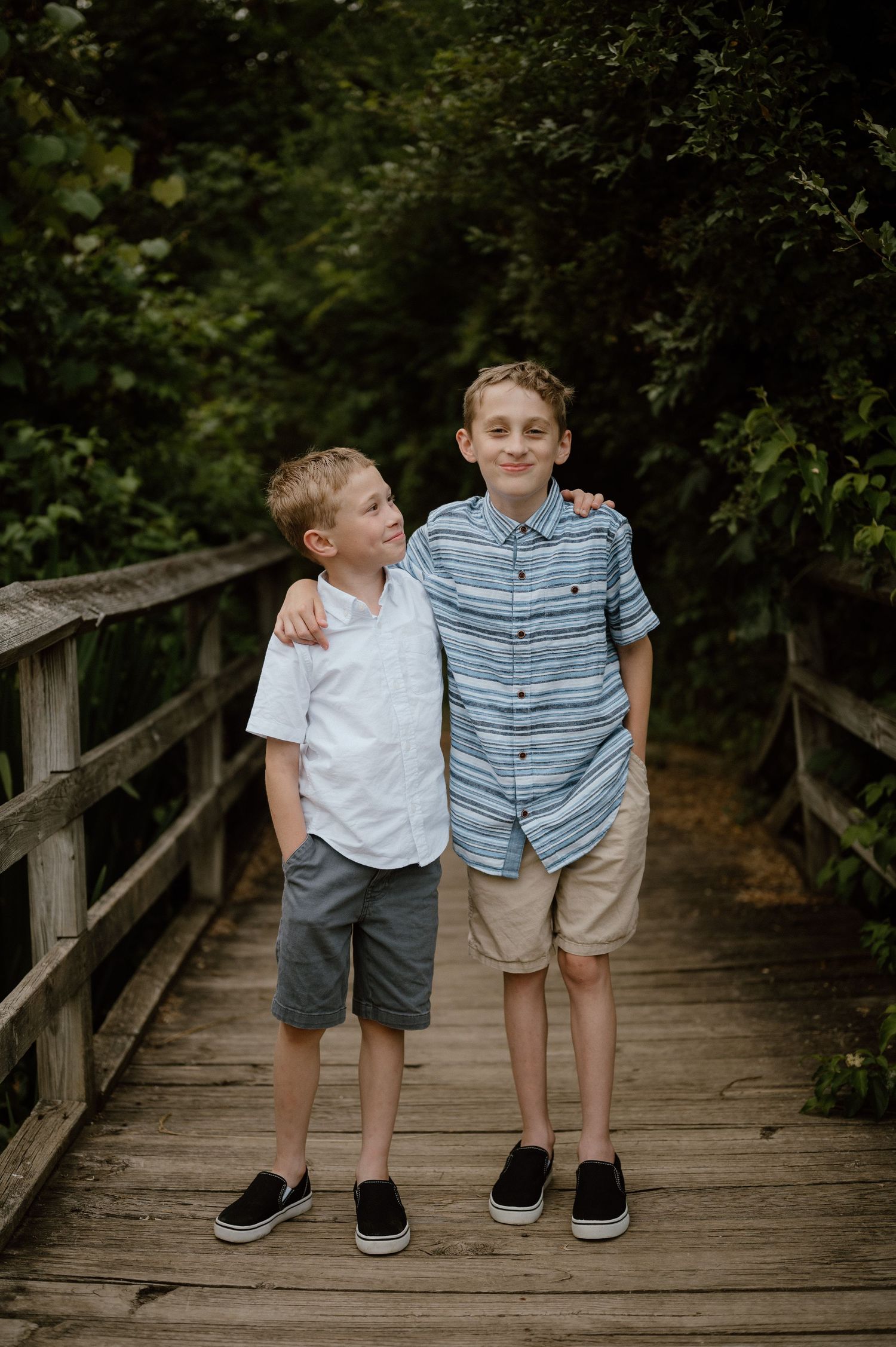 Two children stand together posing for a photo on a wooden bridge surrounded by trees.