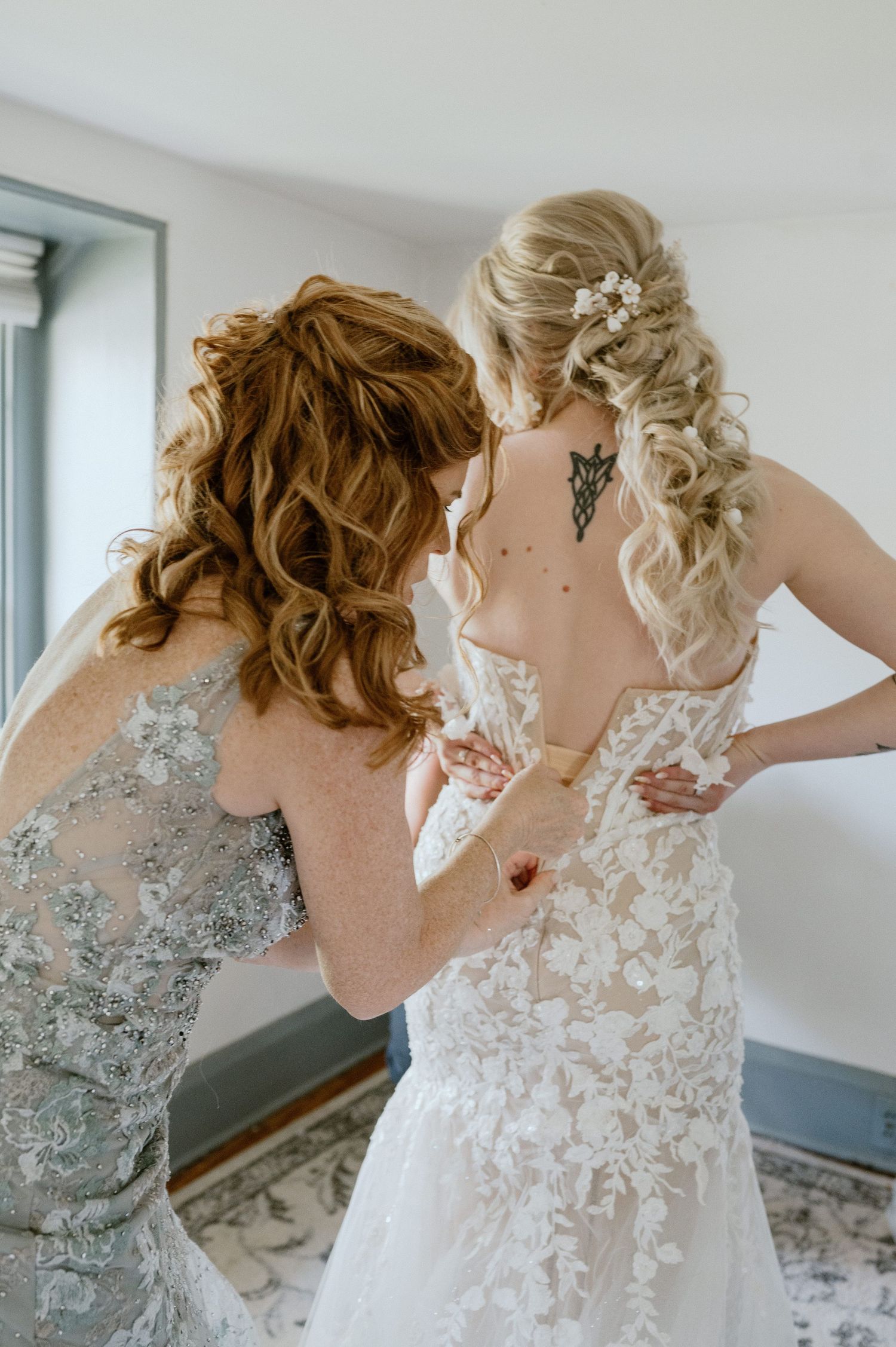 Two women in formal dresses help with buttoning up an ornate white wedding gown.