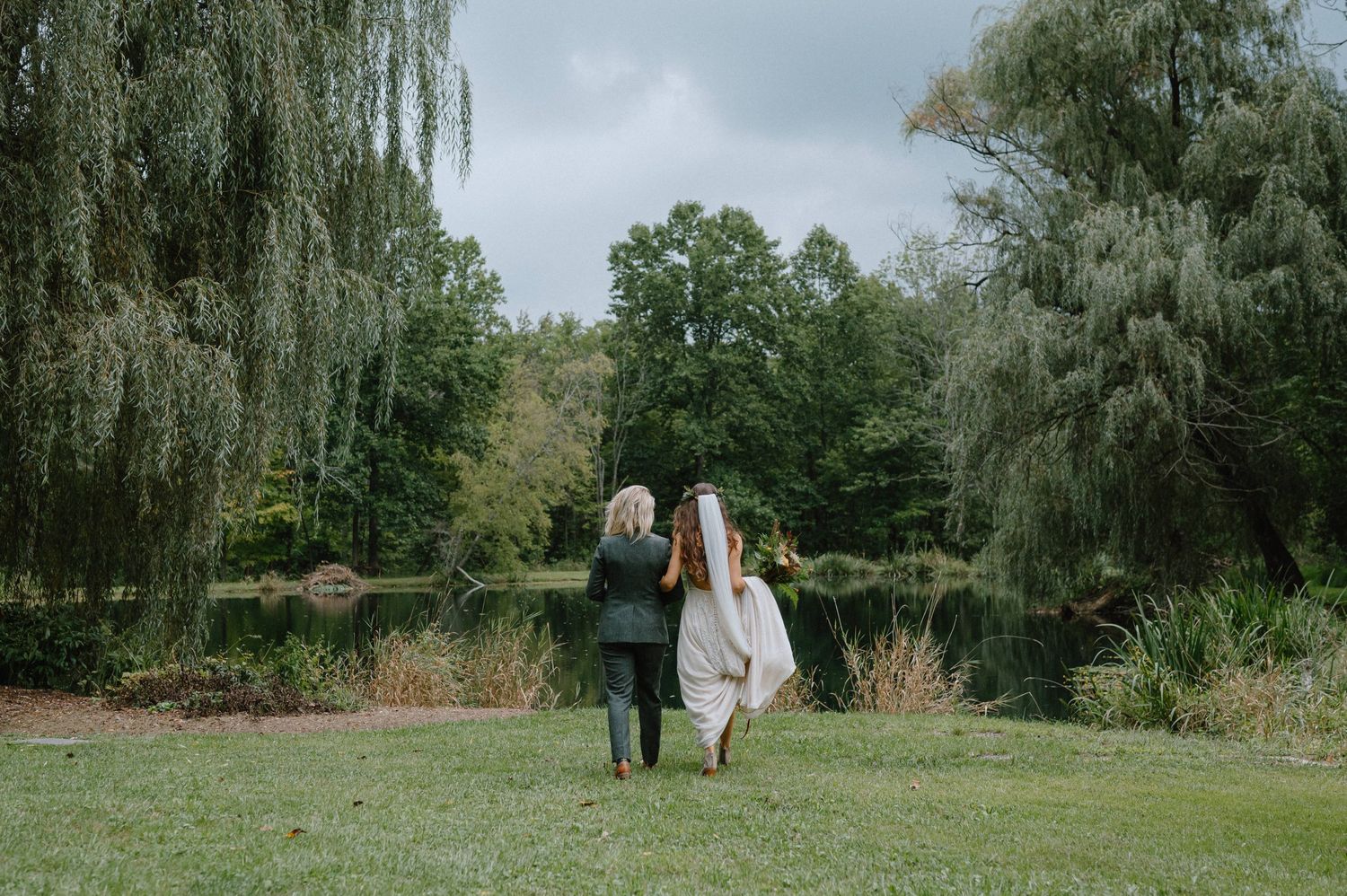 A couple walks through a garden with weeping willow trees on their wedding day.