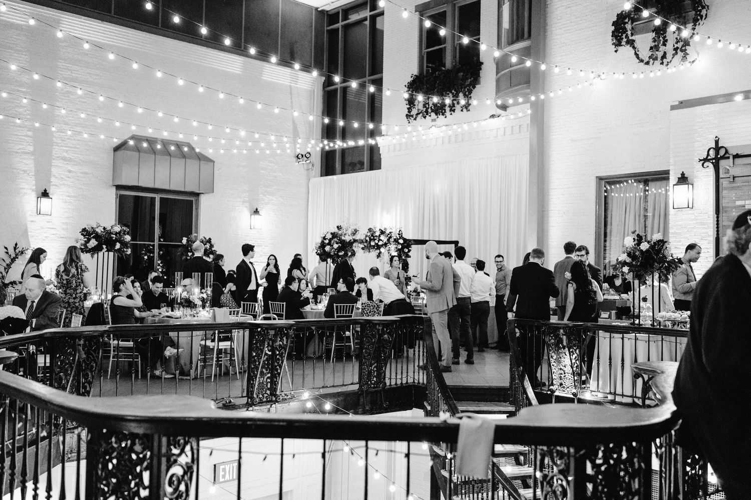 Black and white photo of a wedding reception venue with string lights and guests mingling.