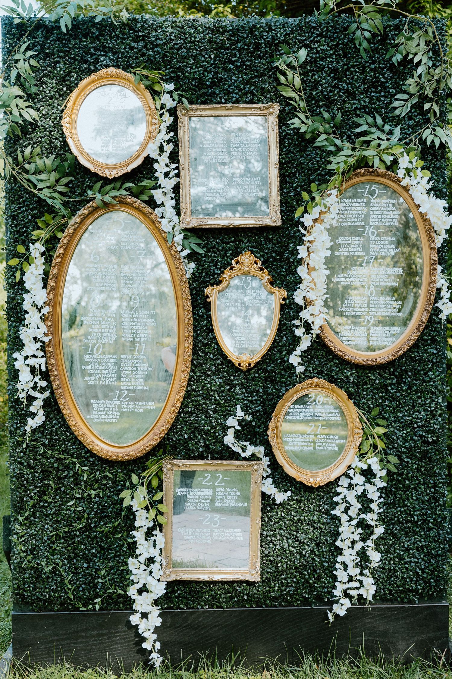 Vintage gold mirrors arranged on a green hedge wall create an elegant wedding seating chart display.