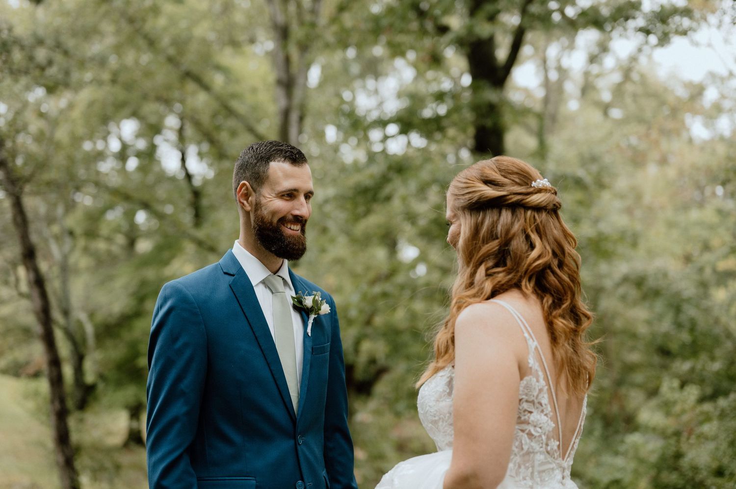 Outdoor wedding moment captured between couple in navy suit and white lace dress under trees.