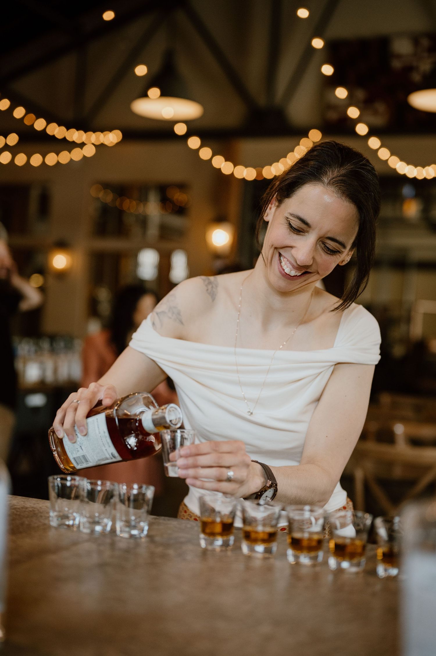 Person in white dress pouring drinks at a bar under warm string lights.