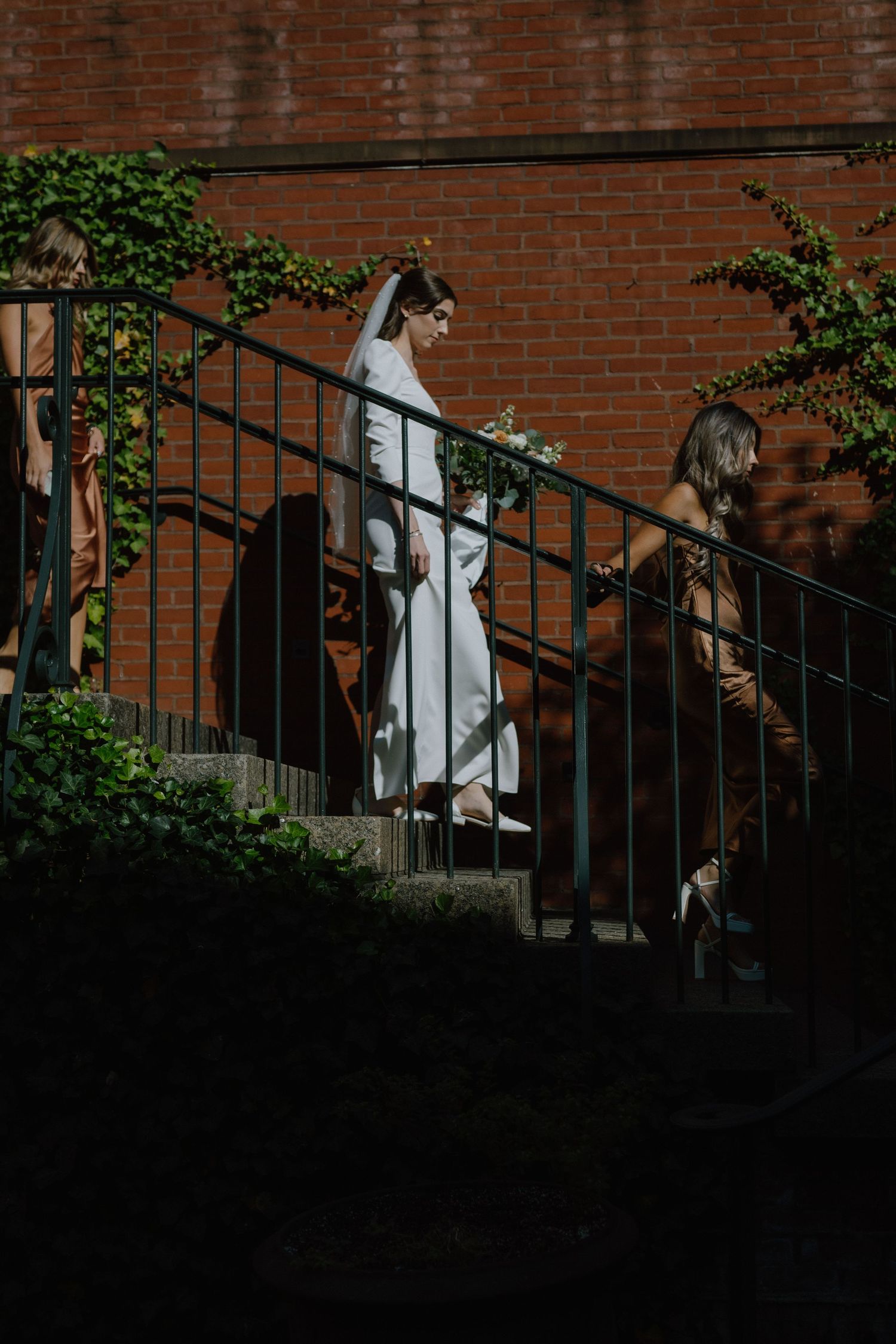 A bride in white descending outdoor stairs with ivy-covered brick walls.