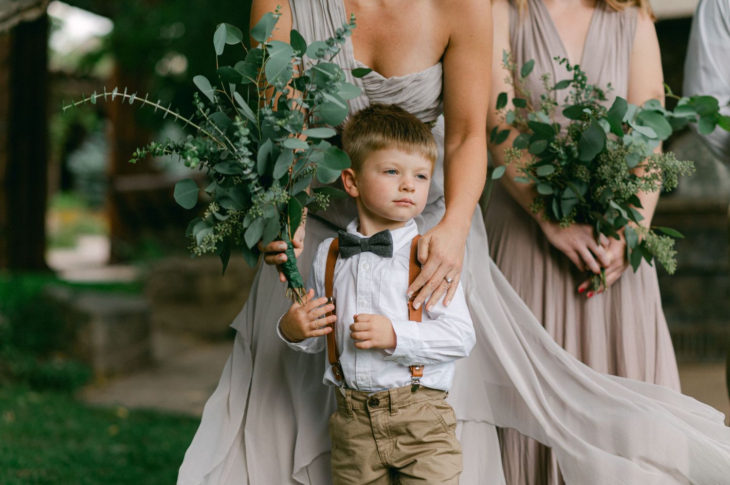A young boy in formal attire stands among wedding attendants holding bouquets of greenery.