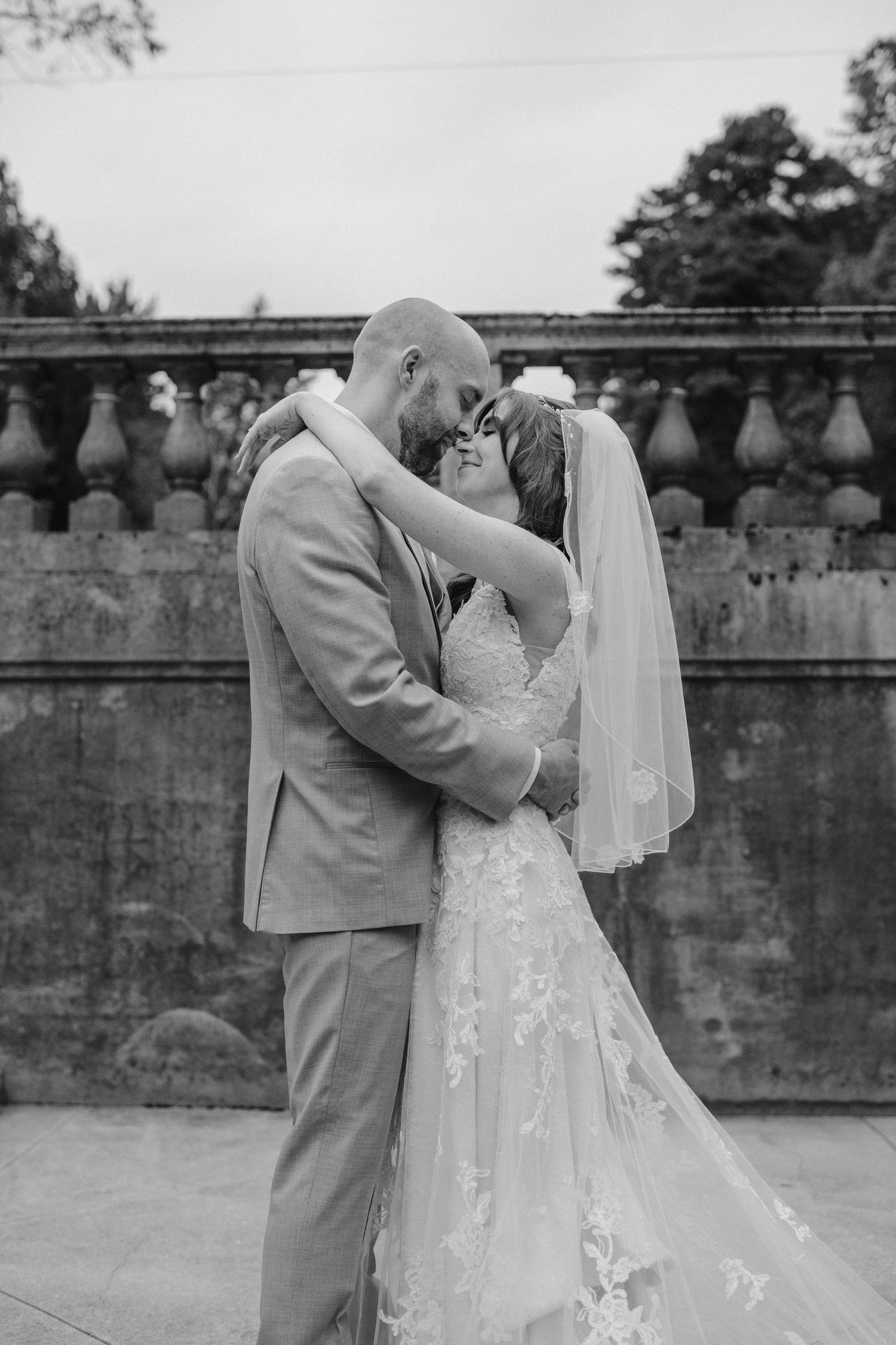 Black and white portrait of newlyweds kissing in front of a stone balustrade.