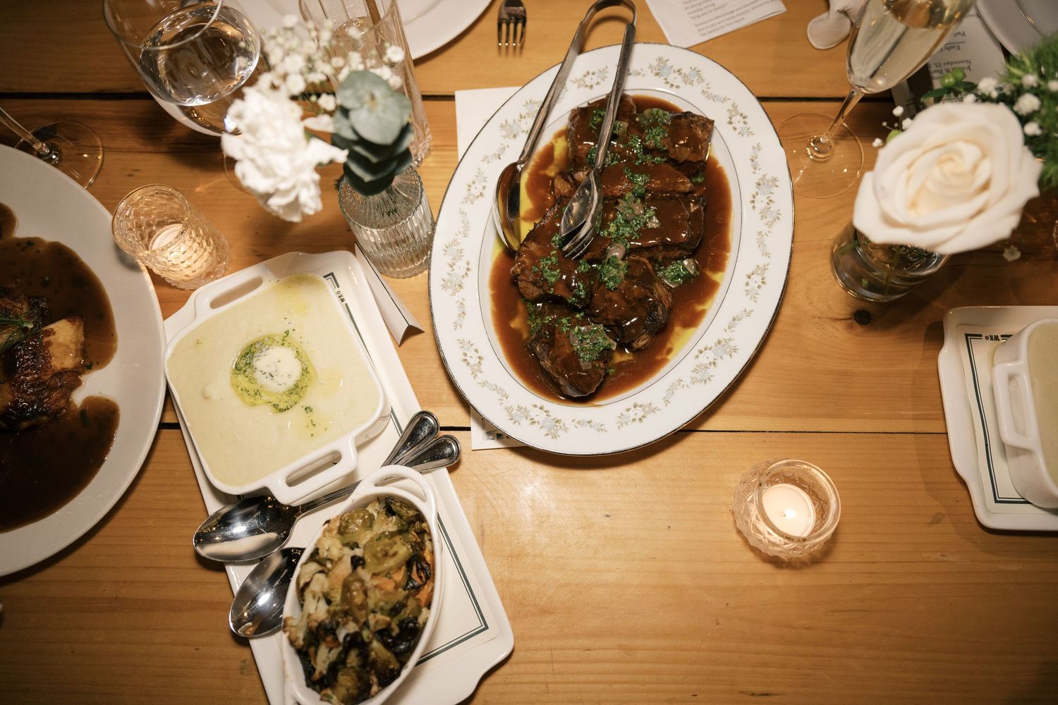 Overhead view of a dinner table with braised meat, side dishes and garnishes served on white plates on a wooden surface.