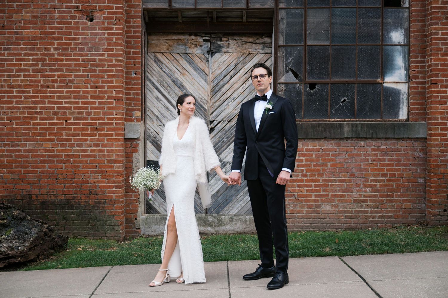 Couple in wedding attire posing against rustic brick wall with wooden door.