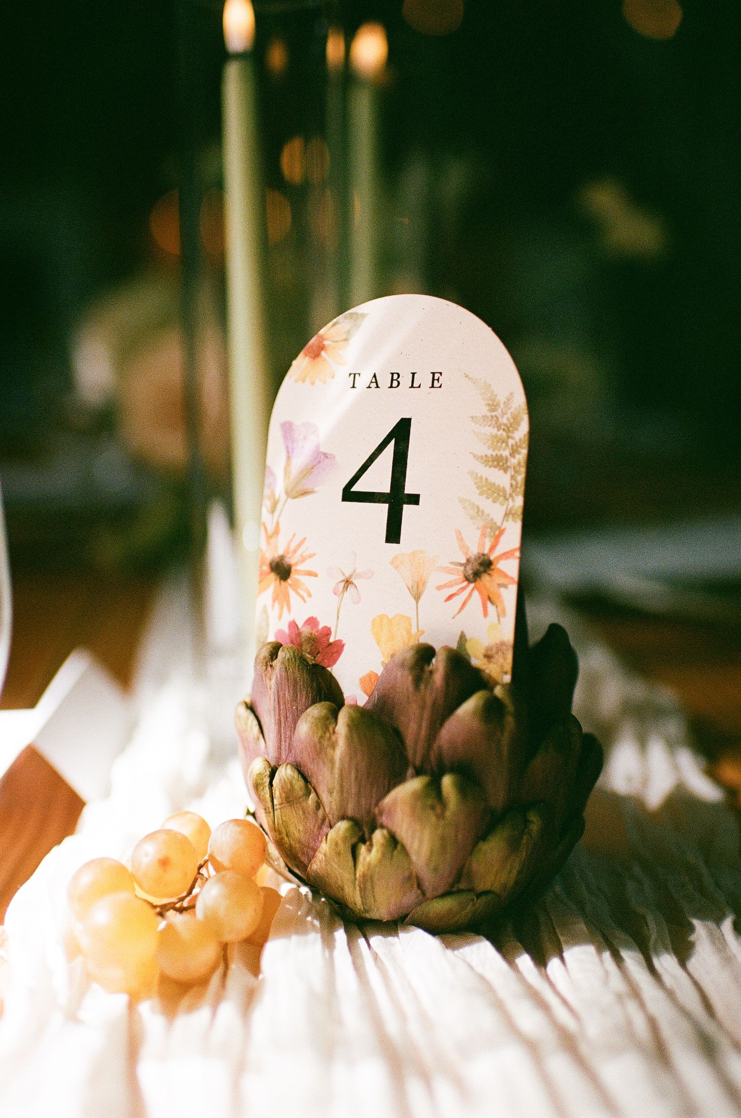 Floral table number card displaying number 4 held in an artichoke centerpiece with grapes at a wedding reception.