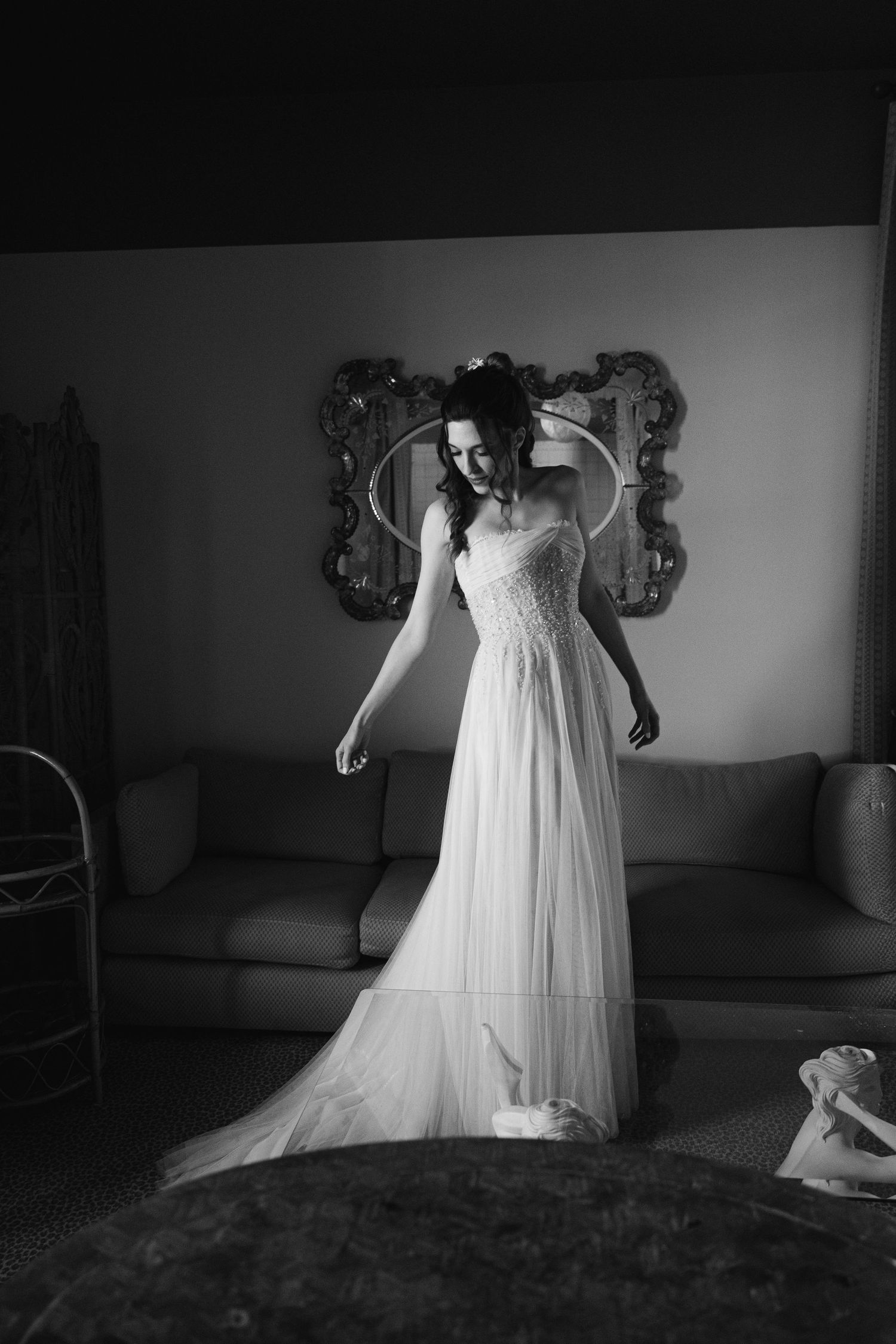 Black and white photo of a bride in a flowing lace wedding gown standing elegantly in a room with an ornate mirror.