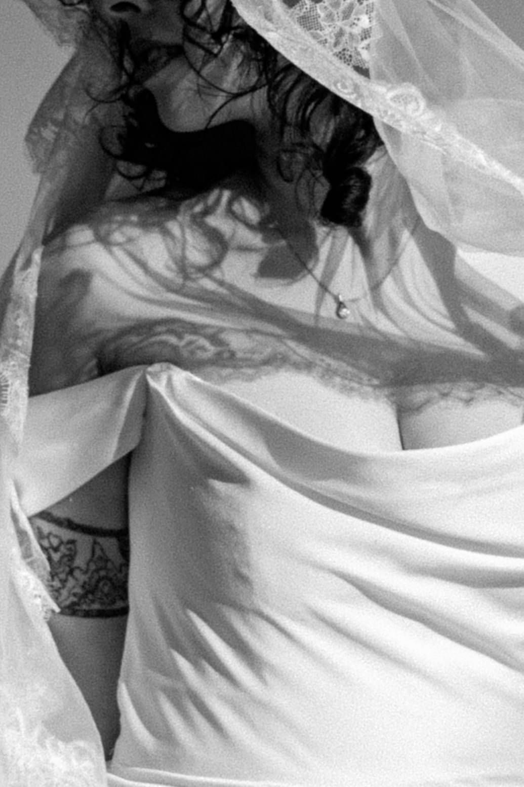 Black and white artistic photo of a woman in a white bridal veil and lingerie with tattoos visible.
