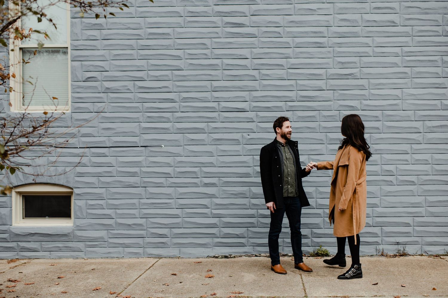 A man and woman holding hands in front of a gray brick wall on a city sidewalk.
