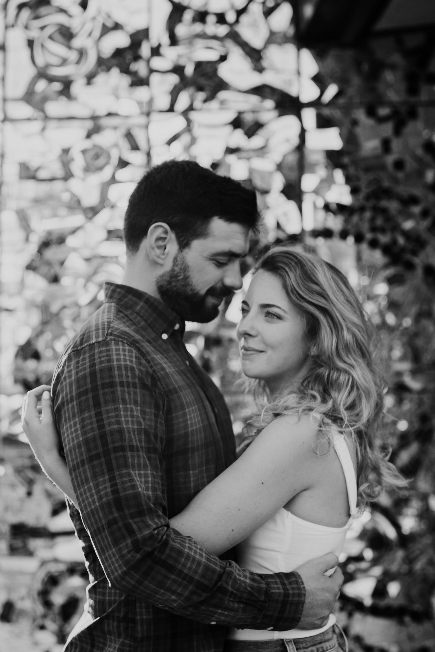 Black and white photo of a couple embracing tenderly, woman smiling softly at camera, decorative mosaic wall behind them.