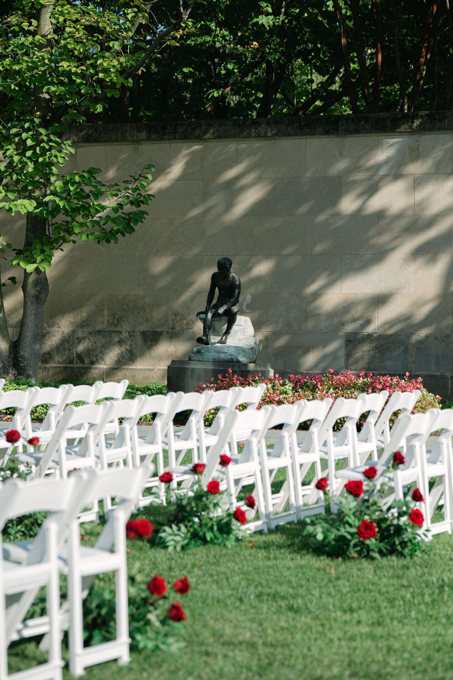 Outdoor wedding ceremony setup with white chairs and red roses against stone wall.